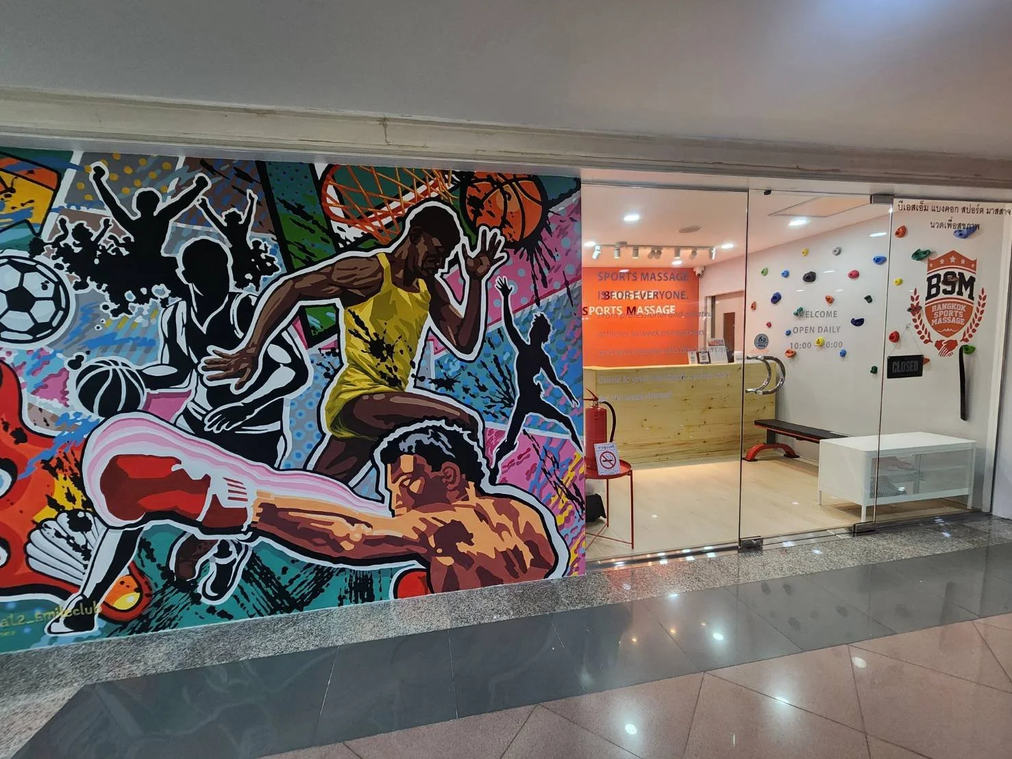 Colorful mural of athletes playing various sports, including basketball, soccer, tennis, and running, near the entrance of a sports massage center in Bangkok.