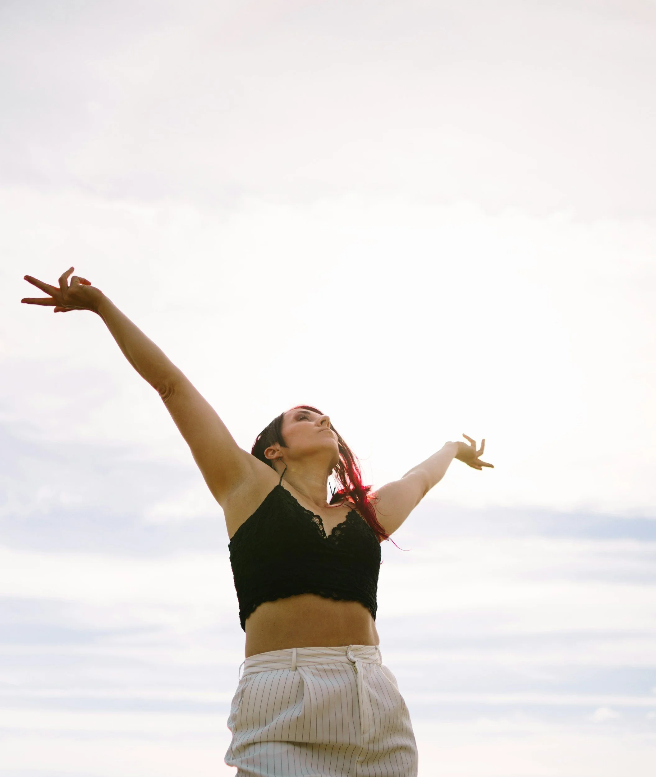 A woman with dark hair and a black lace top stands outdoors against a cloudy sky, with her arms outstretched in a joyful pose.