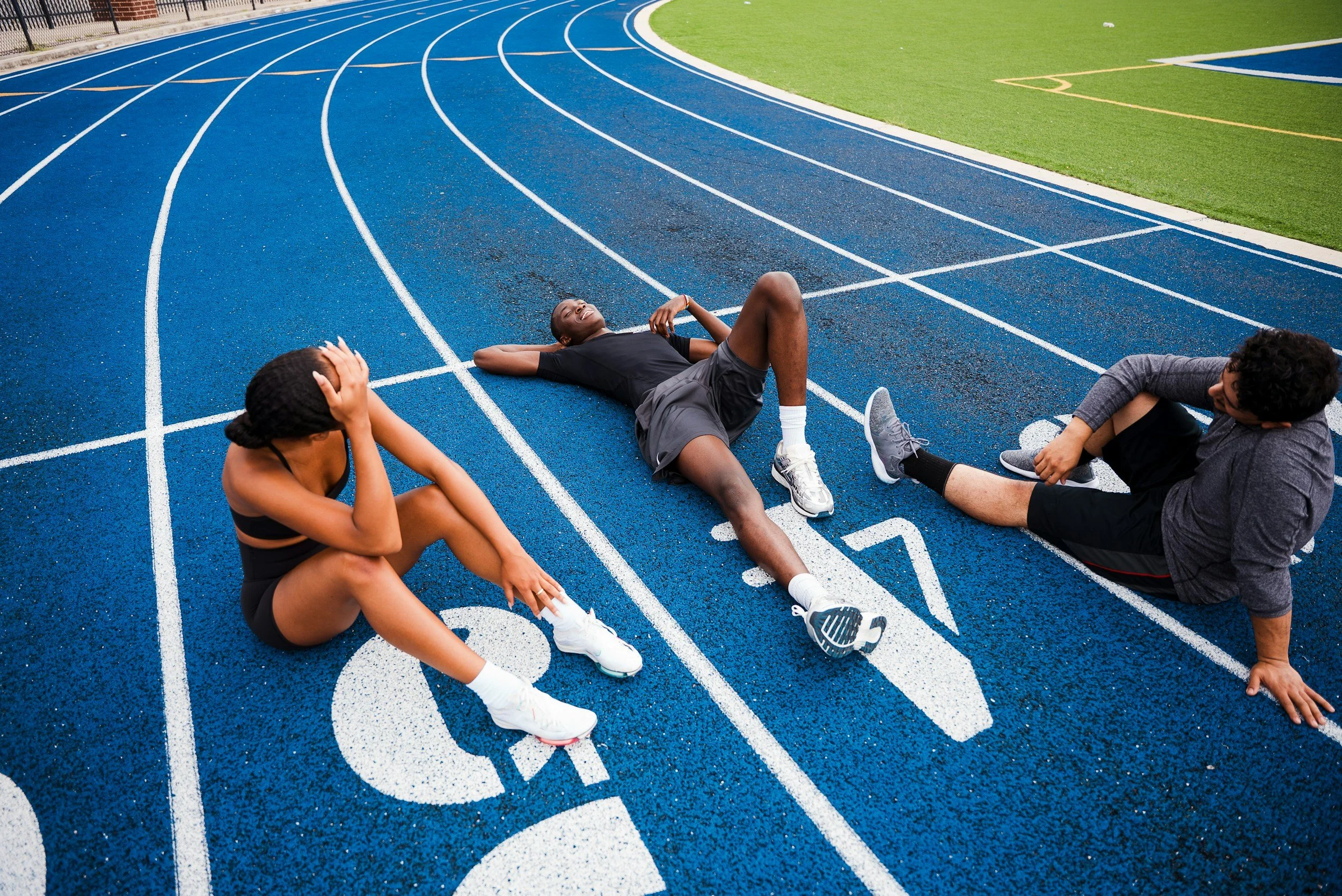 Three athletes sit and lie on a blue running track at a sports stadium, appearing exhausted after a race.