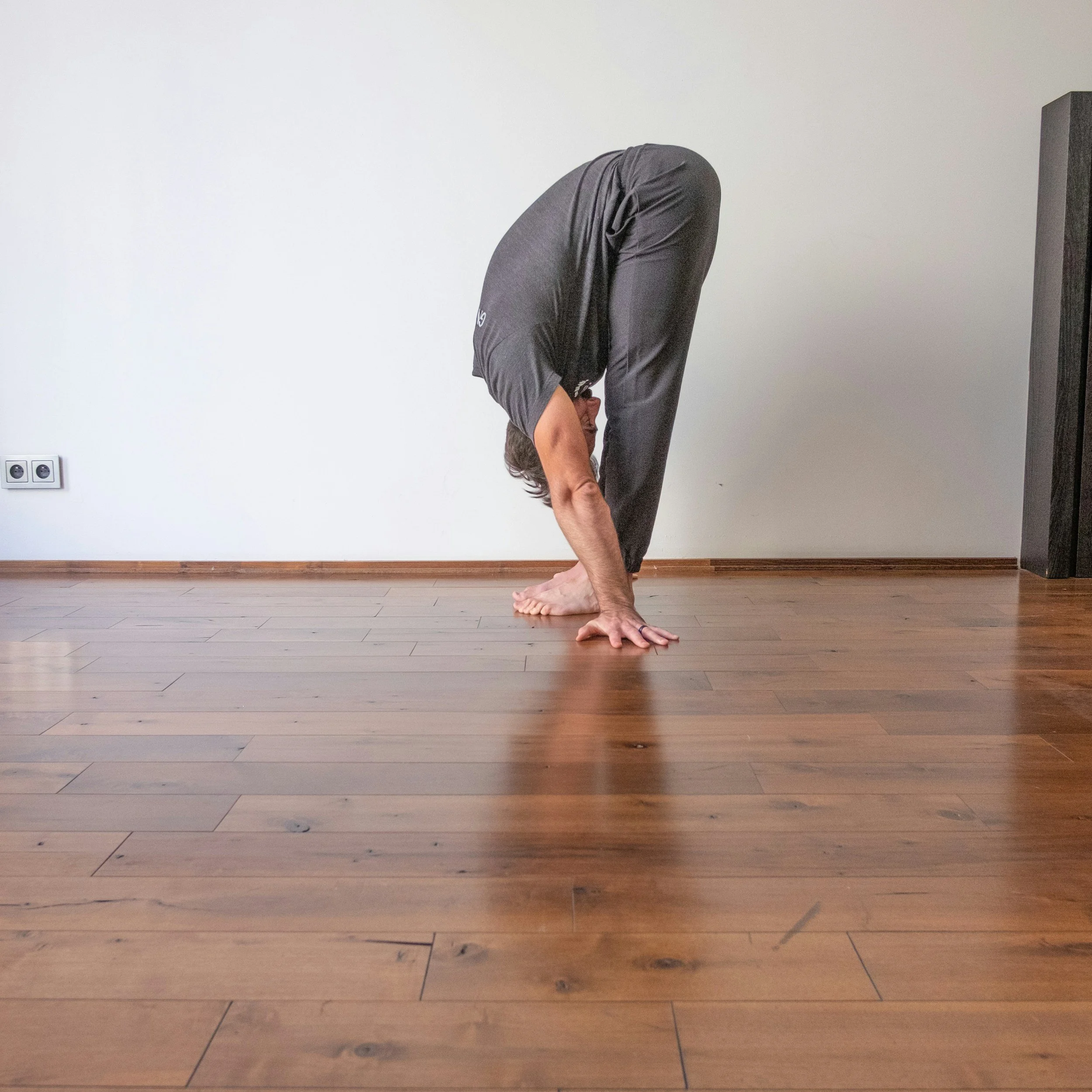 Person performing a forward bend yoga pose in a room with wooden flooring and plain white wall.