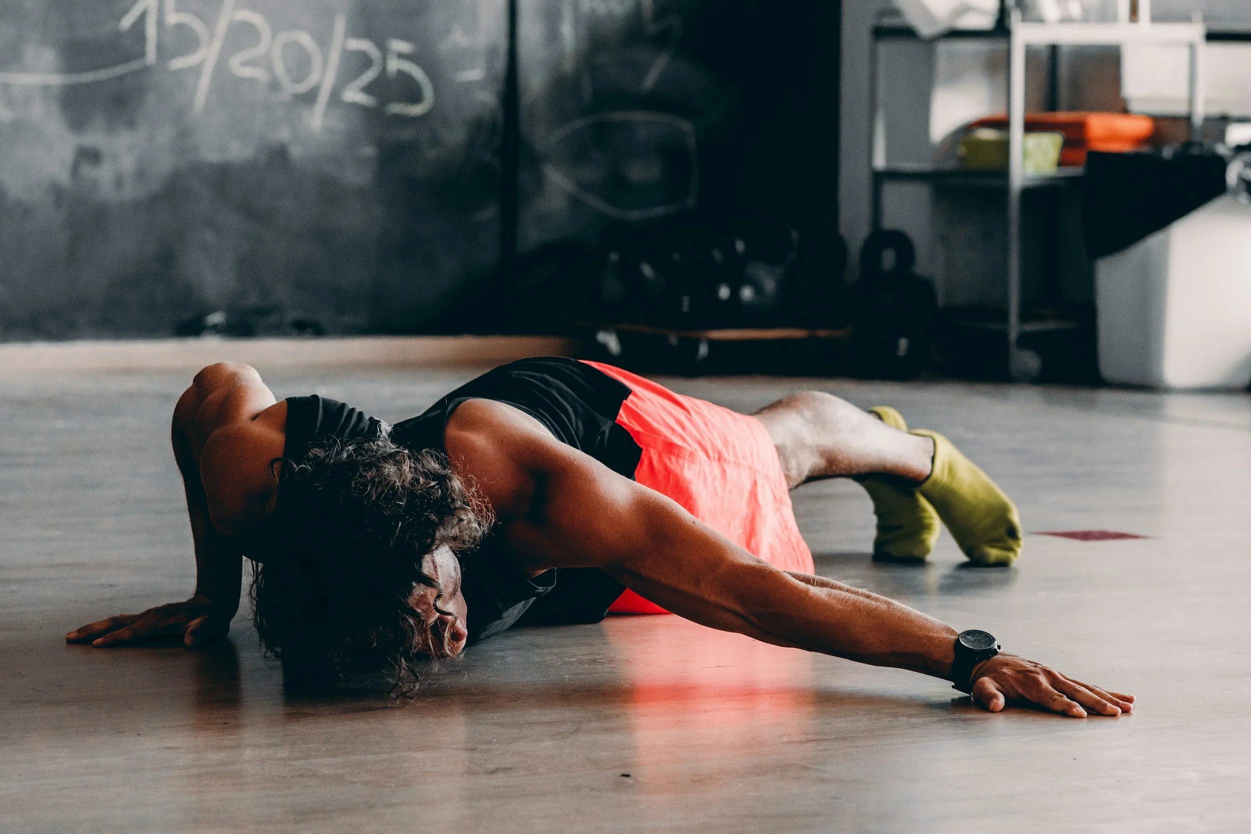 Two women engaging in a stretching or yoga exercise on the floor of a gym, with one woman wearing glasses, black top, and orange shorts, and the other woman in a black top and orange shorts. The background includes a chalkboard with writing, shelves, and workout equipment.