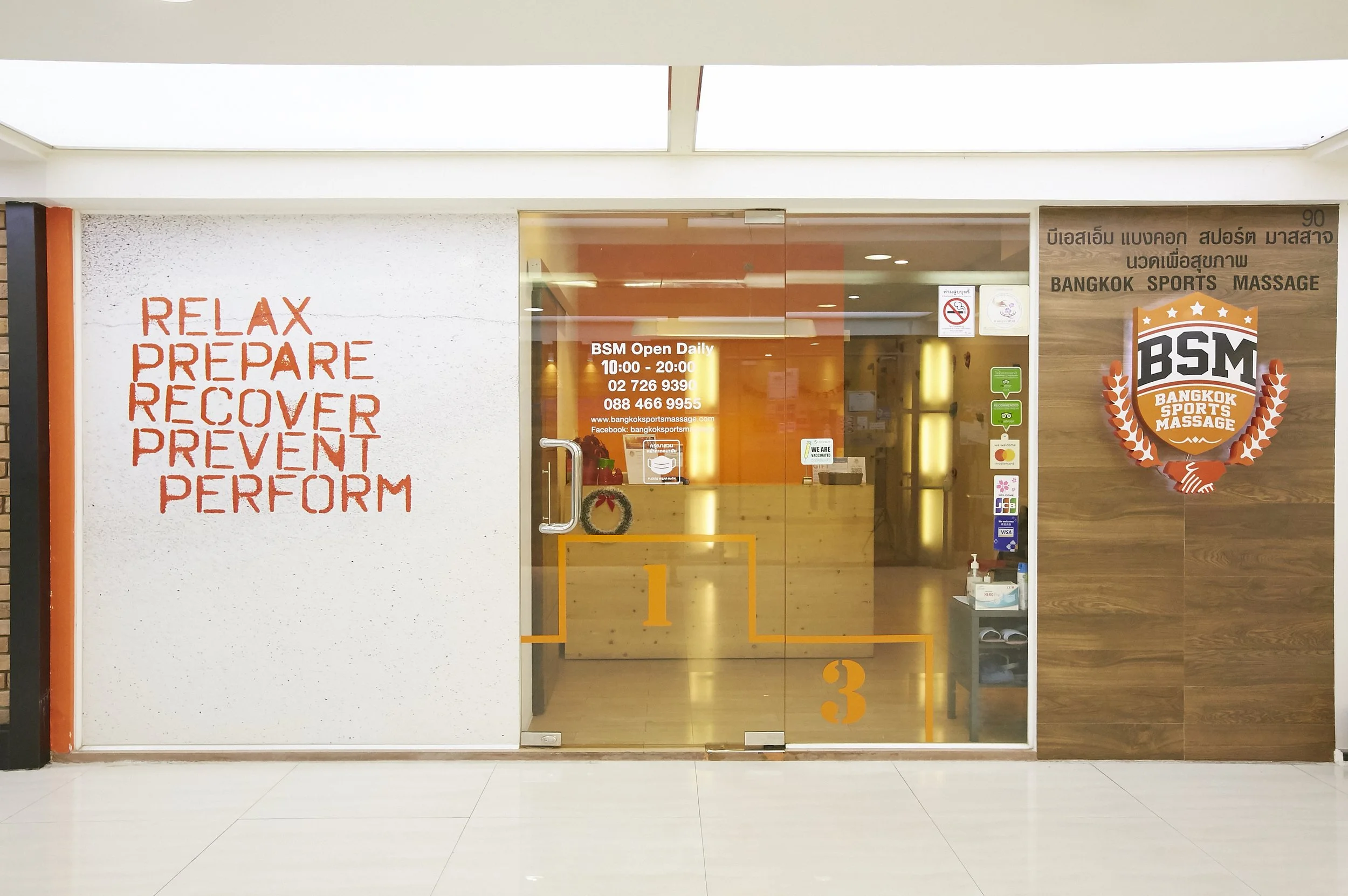 Storefront for Bangkok Sports Massage with glass doors, orange accents, and motivational words like 'Relax,' 'Prepare,' 'Recover,' 'Prevent,' and 'Perform' written in red on the left wall.