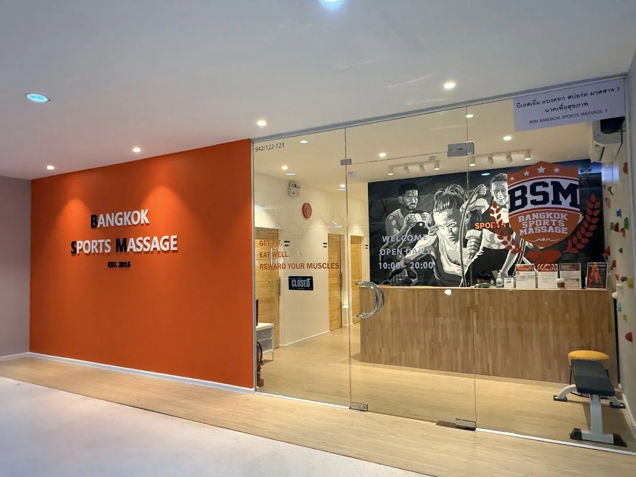 Entrance of a Bangkok Sports Massage center with a glass door, an orange accent wall, and murals of fit athletic individuals.