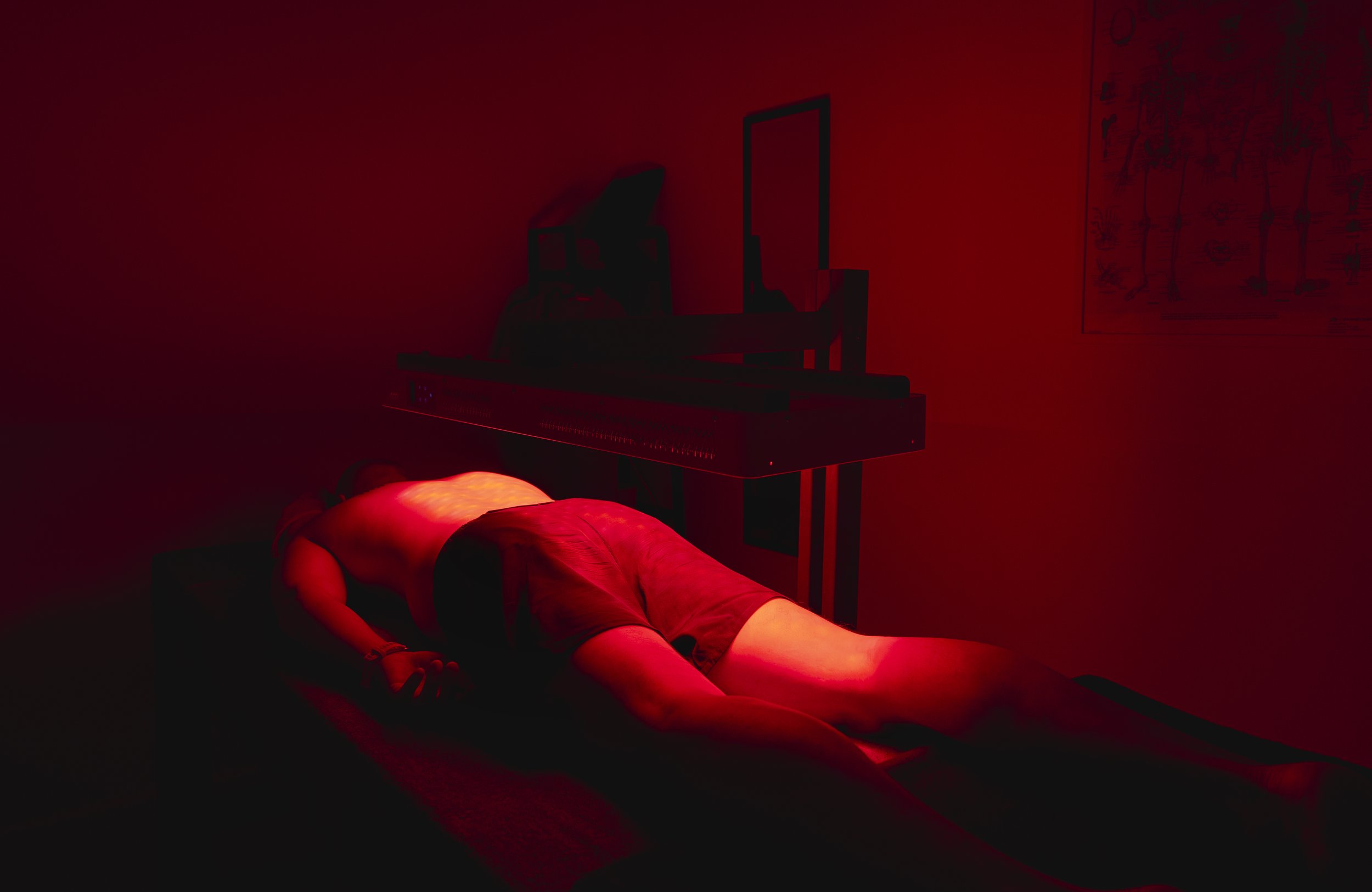 A person lying face down on a massage table in a dimly lit room with red lighting, with a medical or massage device nearby.