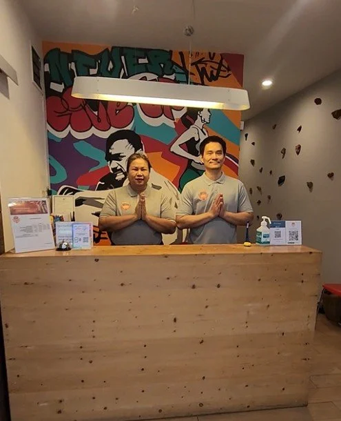 Two staff members standing behind a wooden reception desk with their hands pressed together in a prayer gesture, in a colorful muraled restaurant or cafe. One person has darker skin and the other has lighter skin, both wearing light-colored shirts.