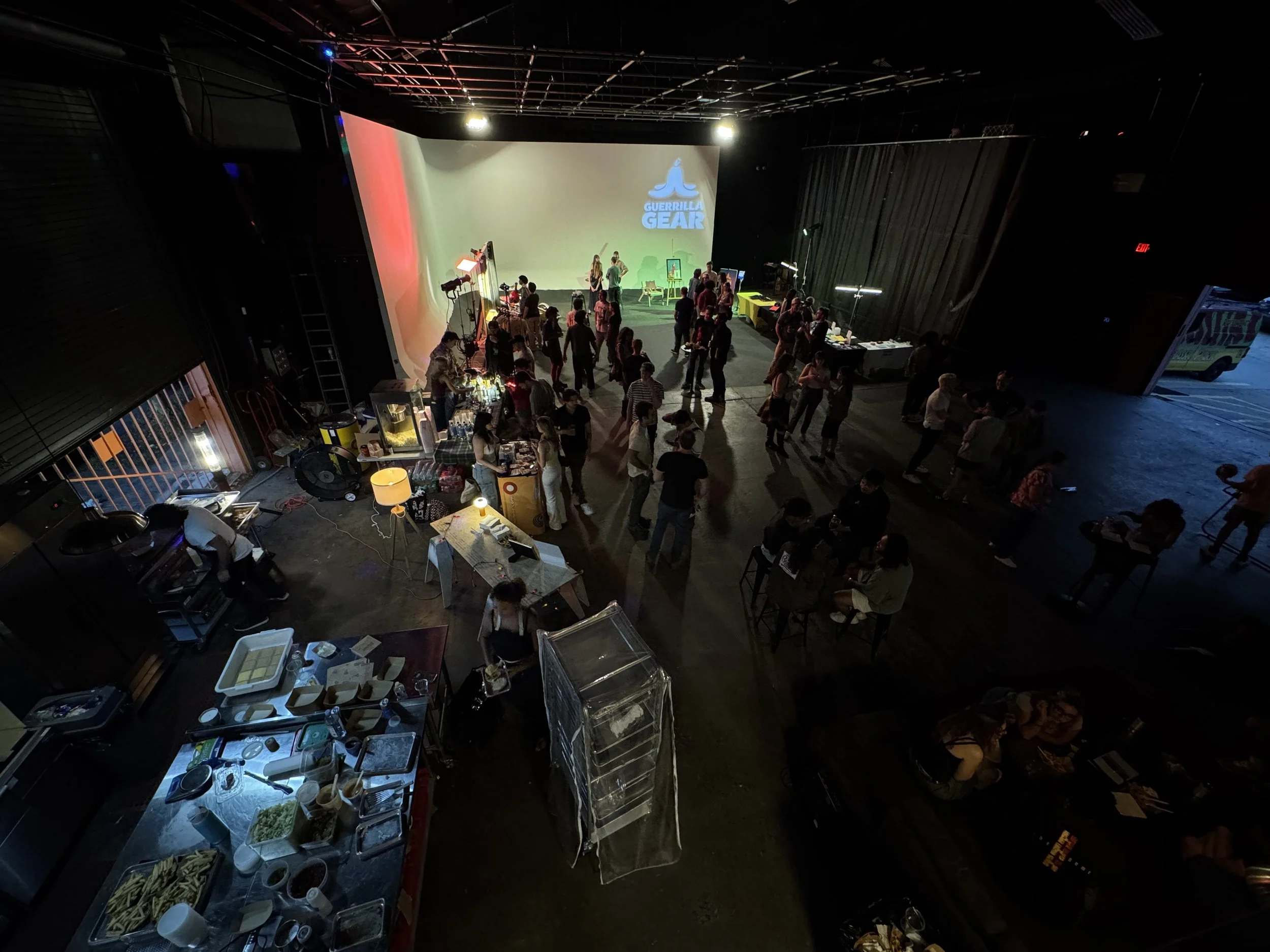 BiRDY. An indoor event with a stage on a large screen displaying a logo for Guerrilla Gear. People are mingling, standing, and sitting at tables. There are food and drink stations with various items, and the lighting is dim with some spotlights.