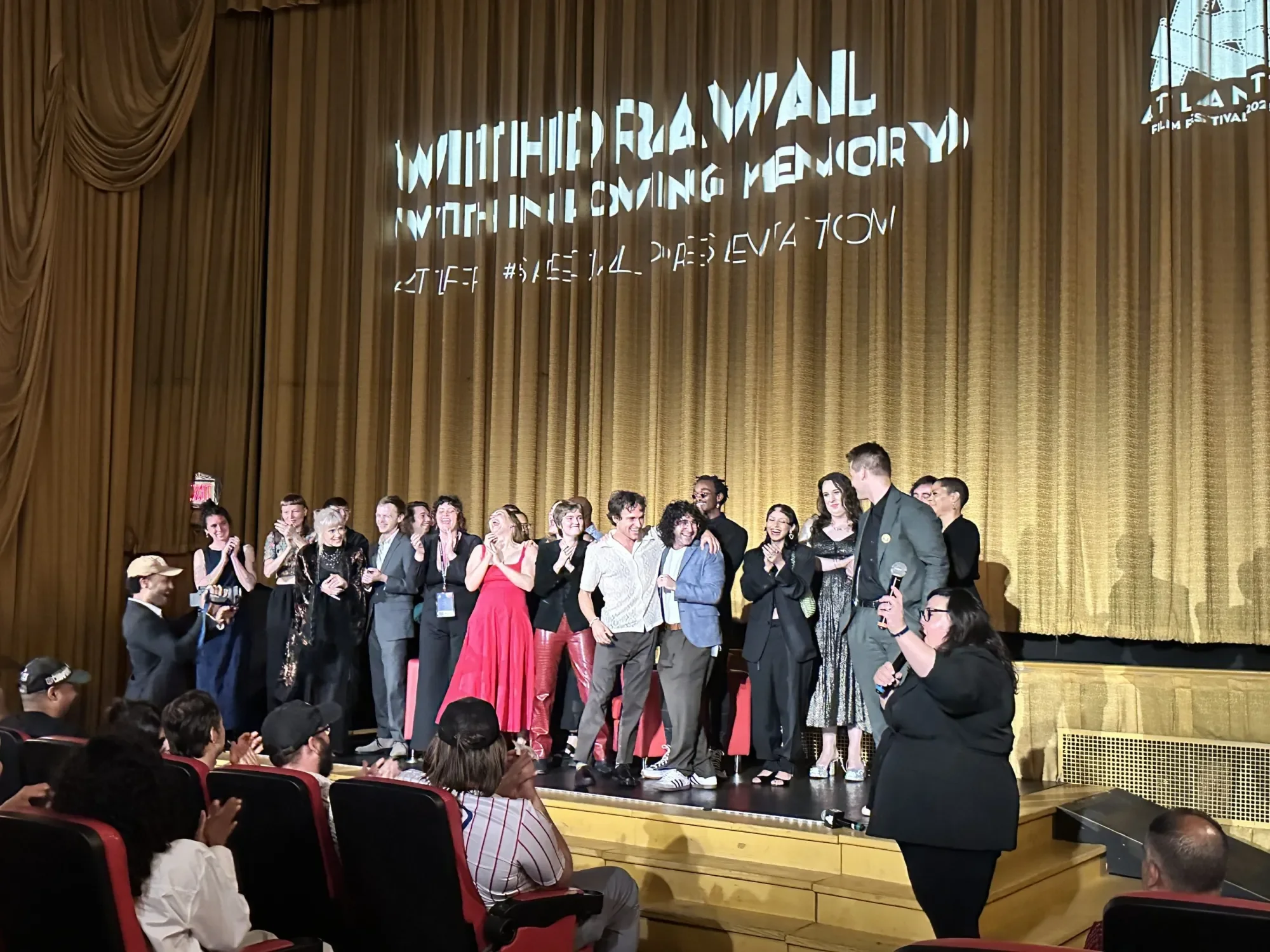 Group of people on stage at the Atlanta Film Festival, some holding microphones, celebrating; audience seated in front. Aaron Strand. Withdrawal Feature Film. Rocco Shapiro. Reel Friends.