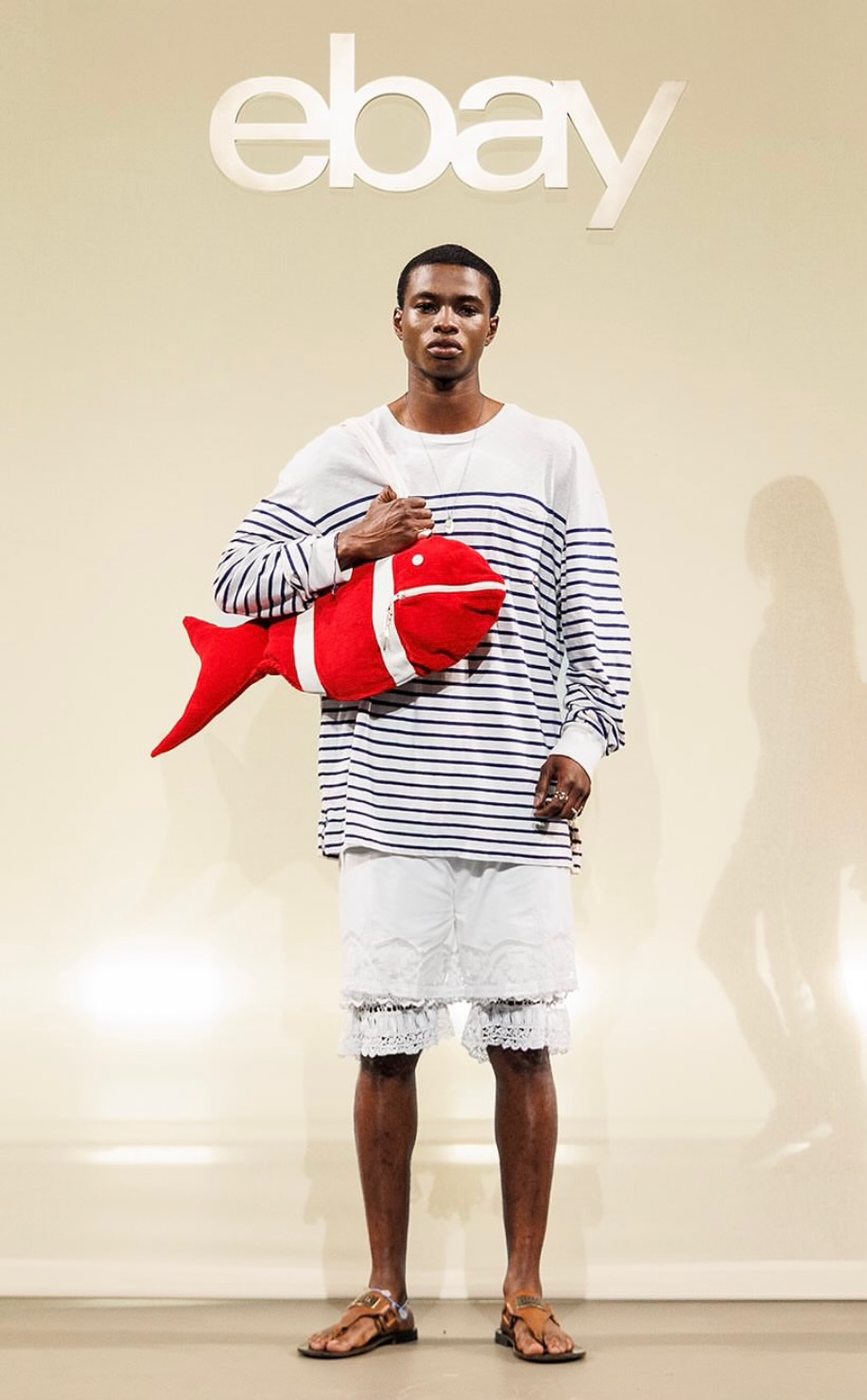 A man standing in front of an eBay logo, holding a plush fish toy, wearing a long-sleeve striped shirt, white shorts, and sandals.