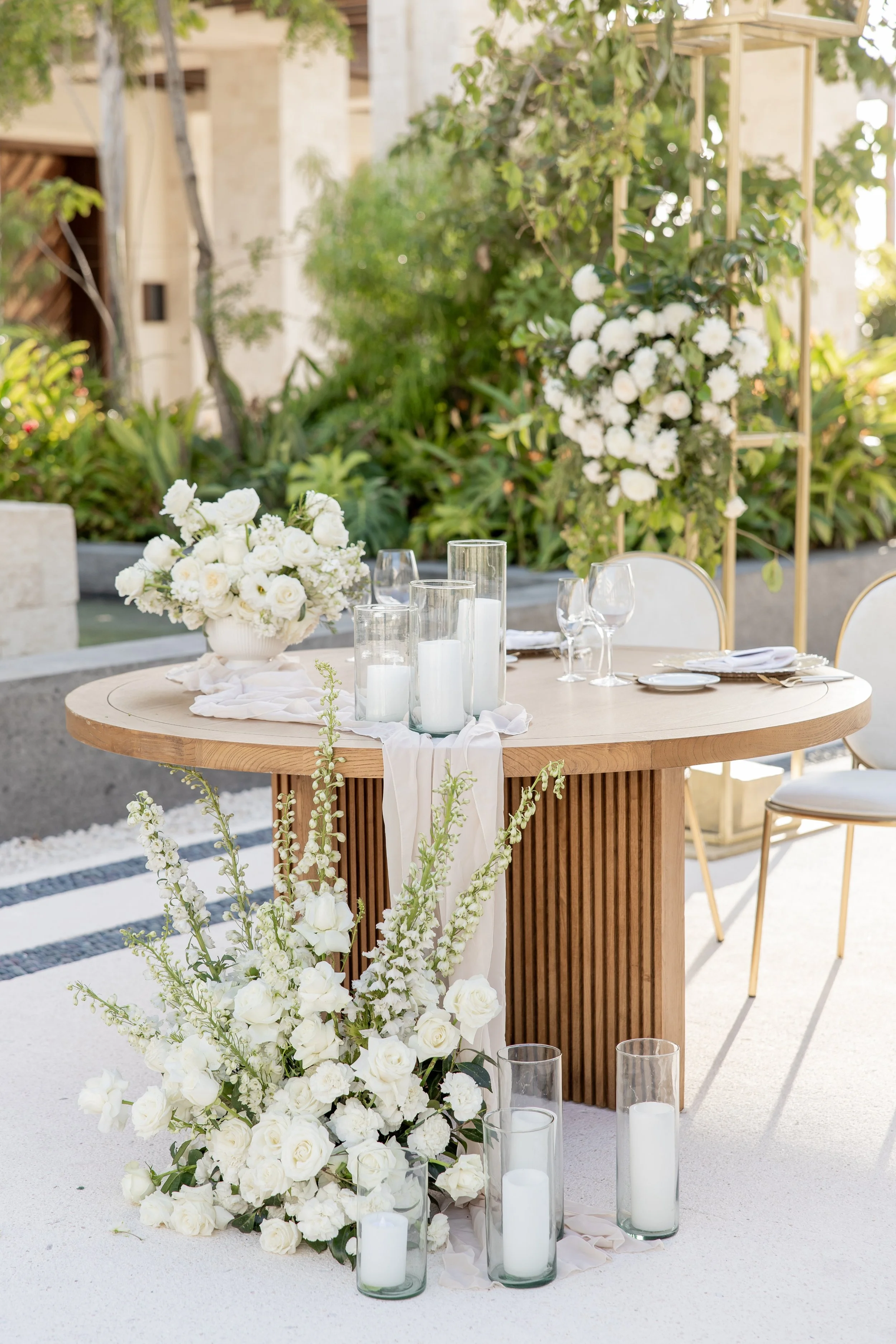 Elegant outdoor wedding table decorated with white flowers, candles, and white tablecloths, set against lush greenery and floral arrangements.