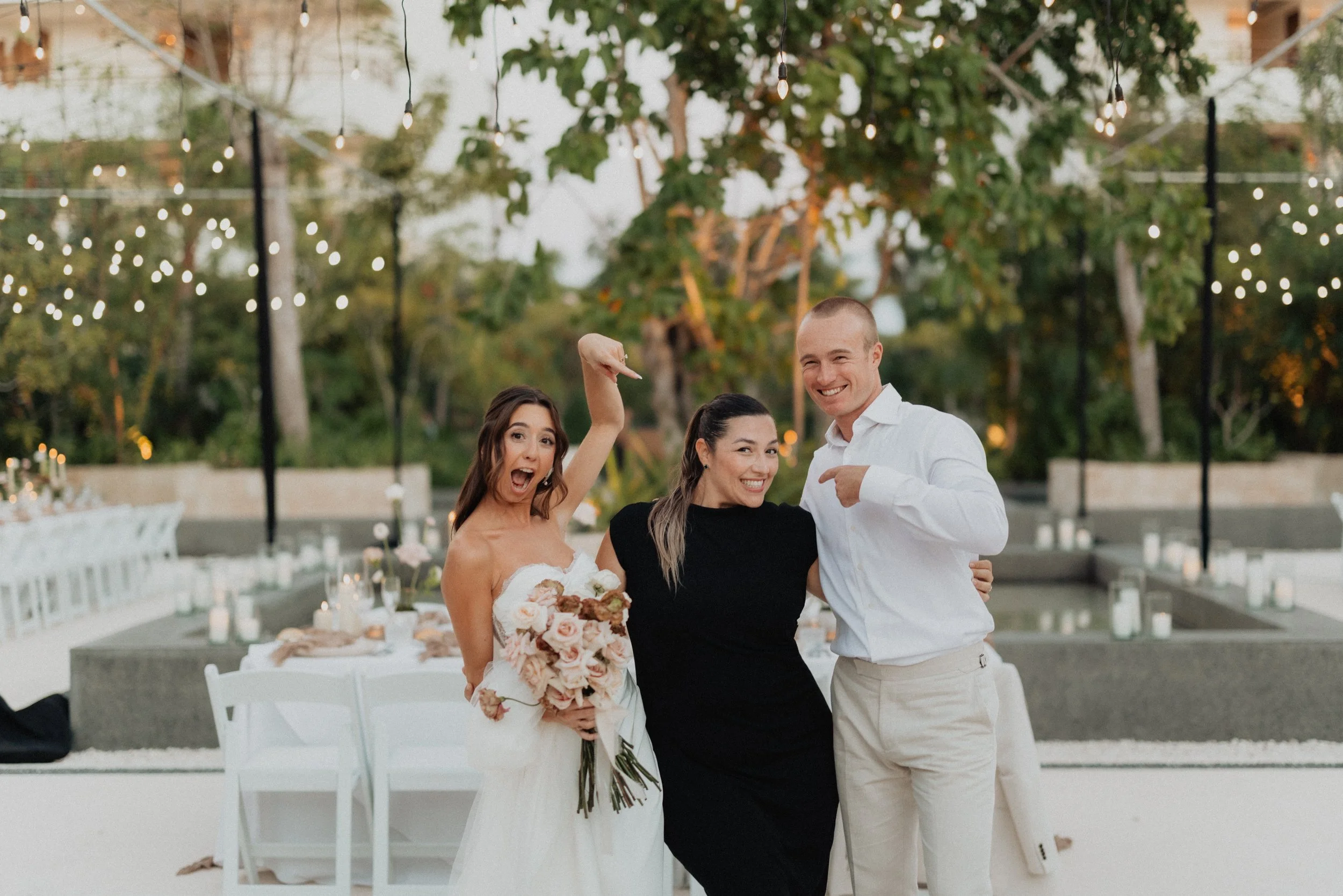 Three people, a bride holding a bouquet and two friends, celebrate at an outdoor wedding reception during the evening with string lights and trees in the background.