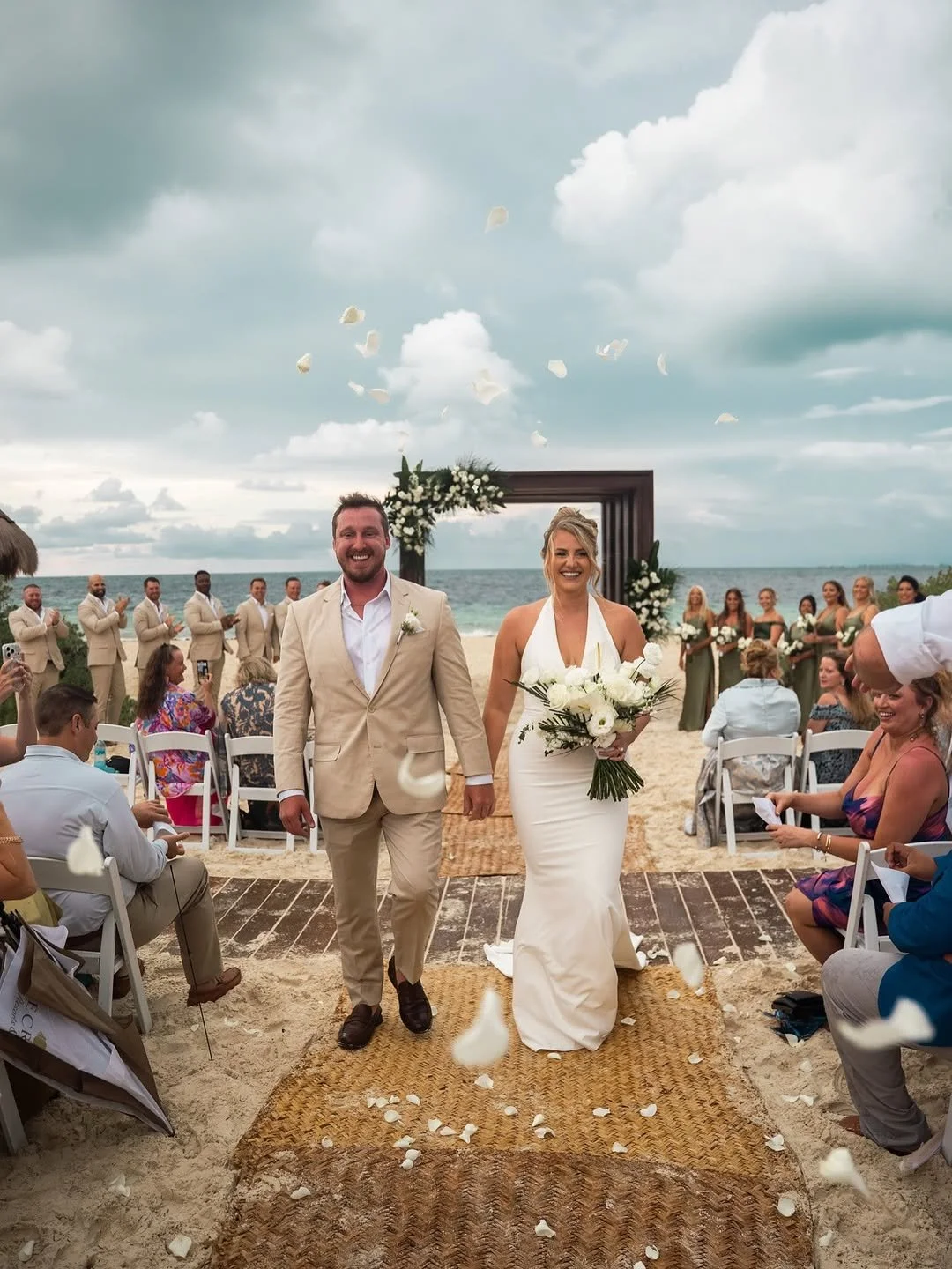A newly married couple walking hand in hand down the aisle at an outdoor beach wedding, with guests seated on either side, a floral arch backdrop, ocean view, and gray clouds in the sky.