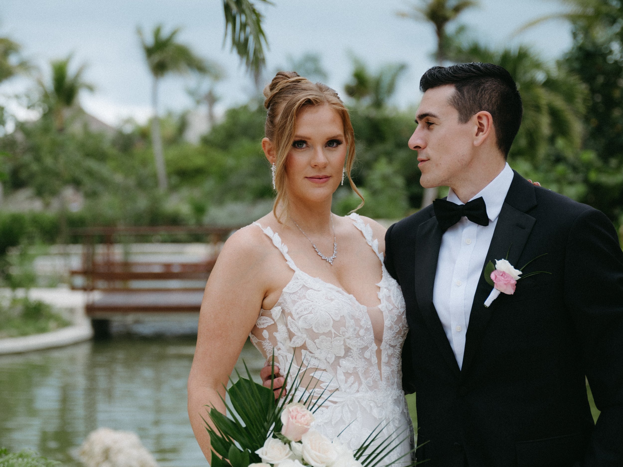 A bride and groom standing close together outdoors, the bride in a white wedding gown holding a bouquet, and the groom in a tuxedo with a pink boutonniere, with lush greenery and a pond in the background.