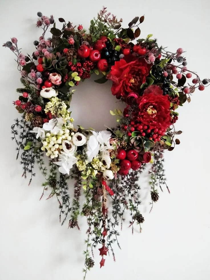 A heart-shaped Christmas wreath decorated with red and white flowers, berries, and greenery, hanging against a white wall.
