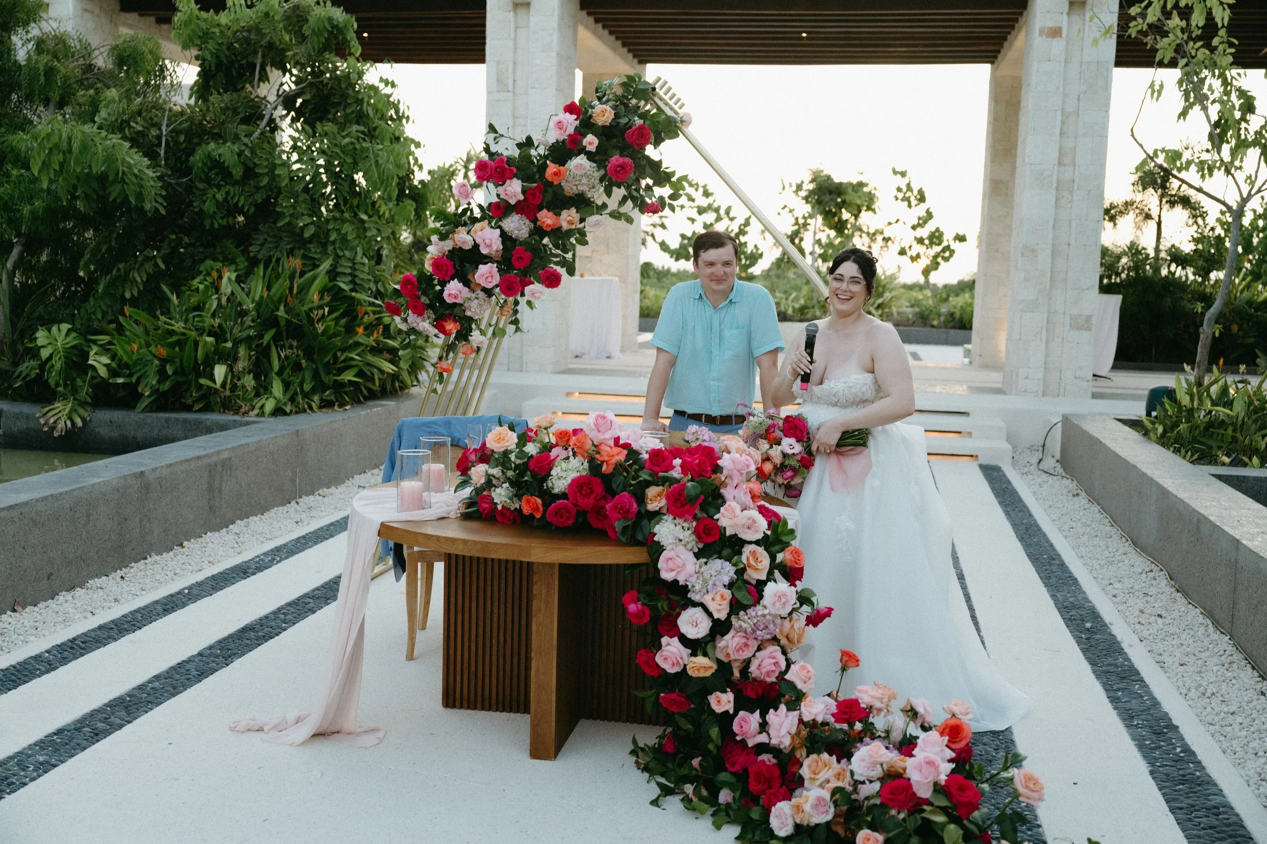Bride and groom standing behind a table with floral arrangements at a wedding ceremony in an outdoor setting.