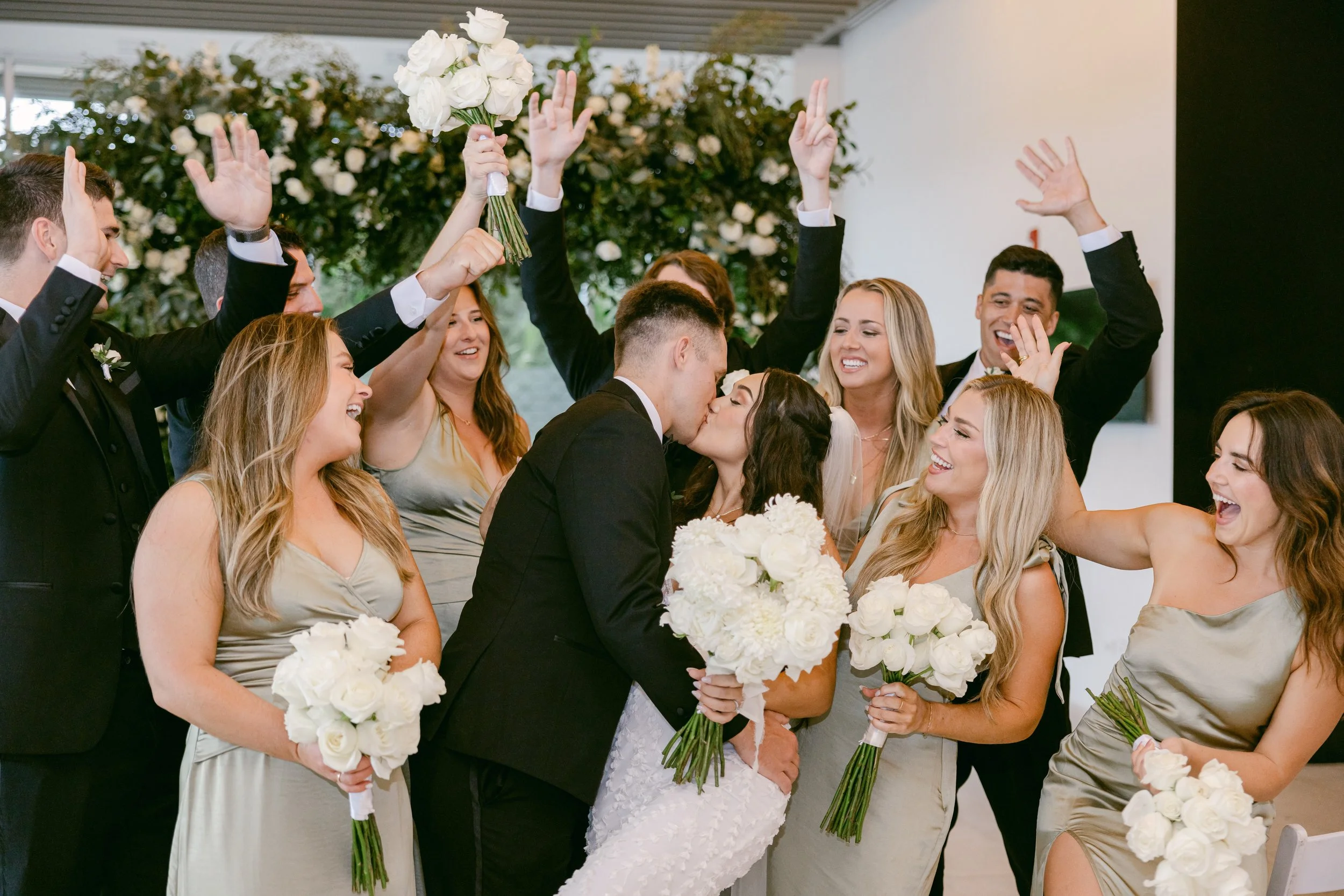 A wedding celebration with the bride and groom kissing surrounded by friends and family holding white bouquets, some raising hands and throwing flowers in the air.
