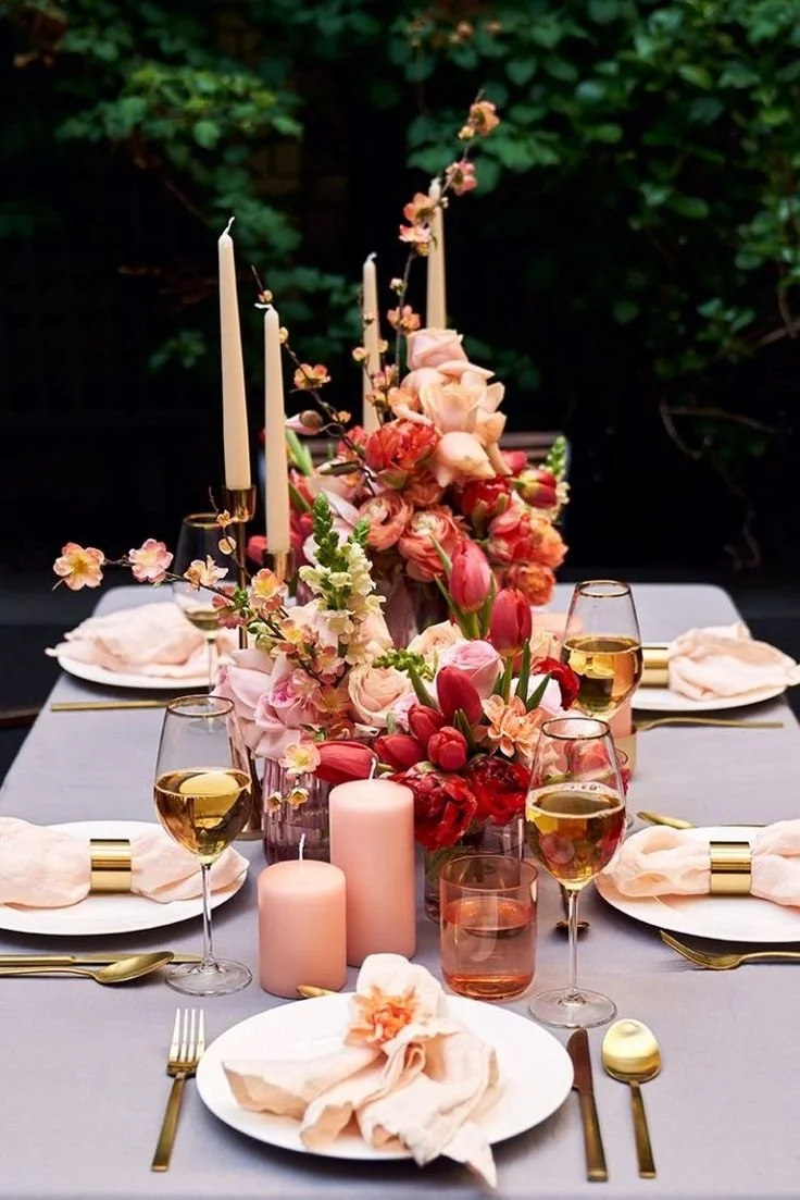 Elegant outdoor table setting with pink flowers, candles, white plates, gold cutlery, and glasses of white wine on a gray tablecloth.