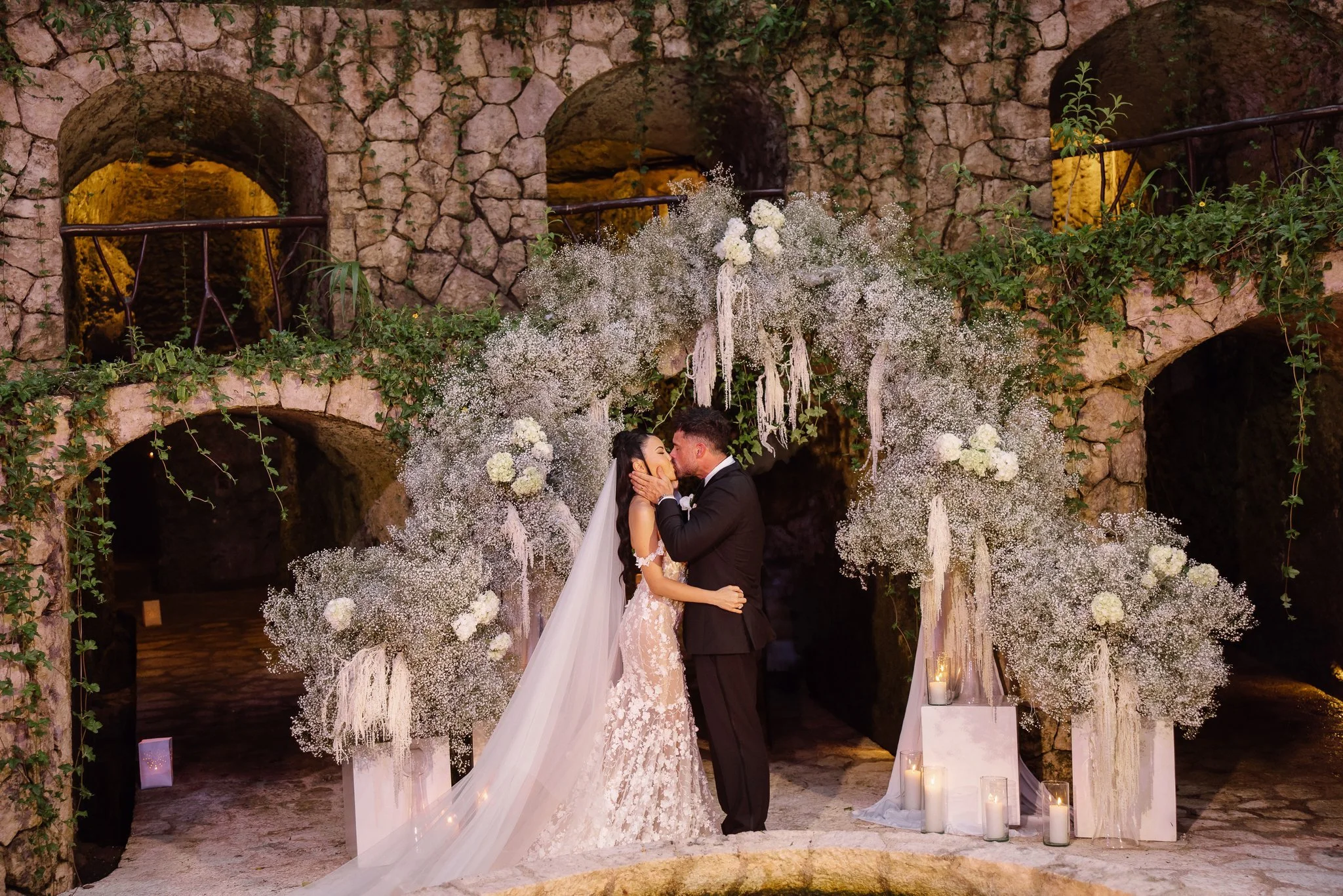 A bride and groom kissing during their wedding ceremony at an outdoor arched stone venue, decorated with white flowers and candles.