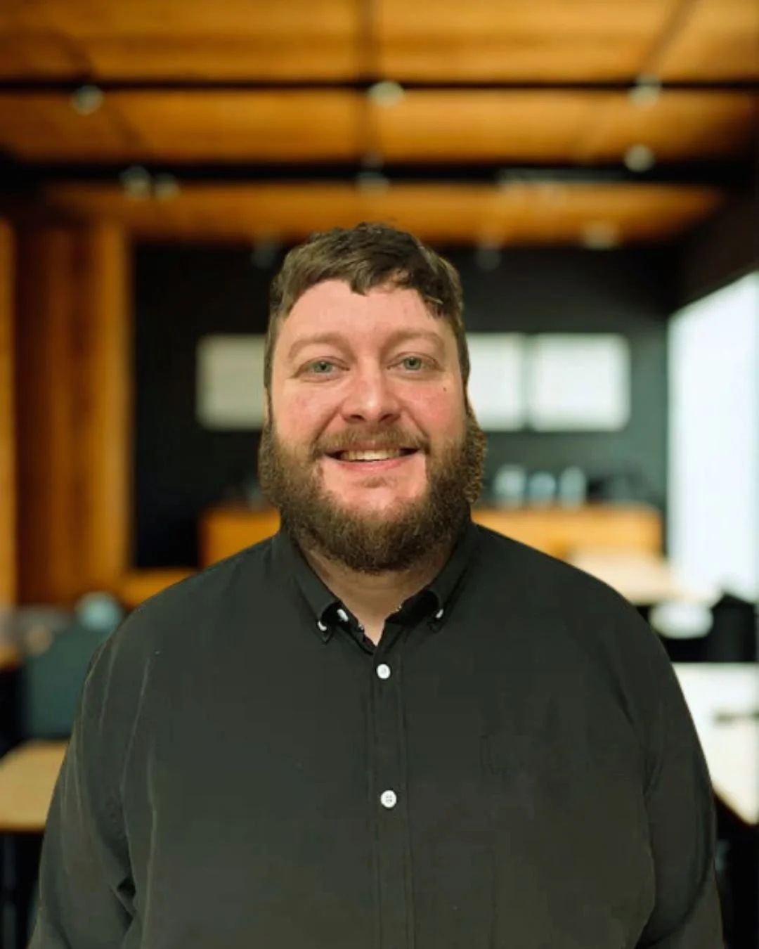 A man with a beard and short hair smiling, wearing a black button-up shirt, standing in a modern office with wooden ceiling panels and blurred background.