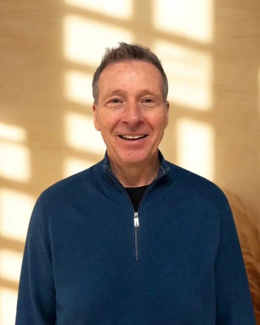 A middle-aged man with short brown hair smiling, wearing a blue quarter-zip sweater, standing indoors with sunlight casting a pattern of shadows on the beige wall behind him.
