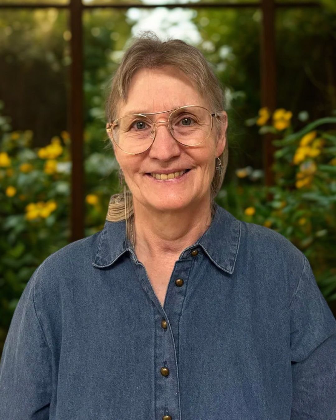 A smiling woman with glasses and earrings in a denim shirt standing outdoors with yellow flowers and greenery in the background.
