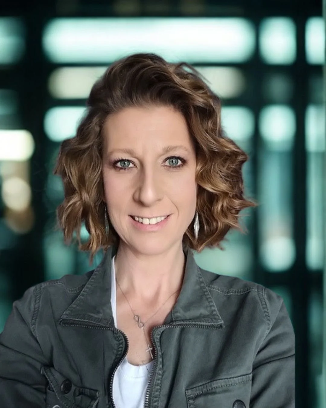 Portrait of a woman with shoulder-length curly hair, wearing a dark gray jacket and white top, smiling in front of a blurred background of windows.