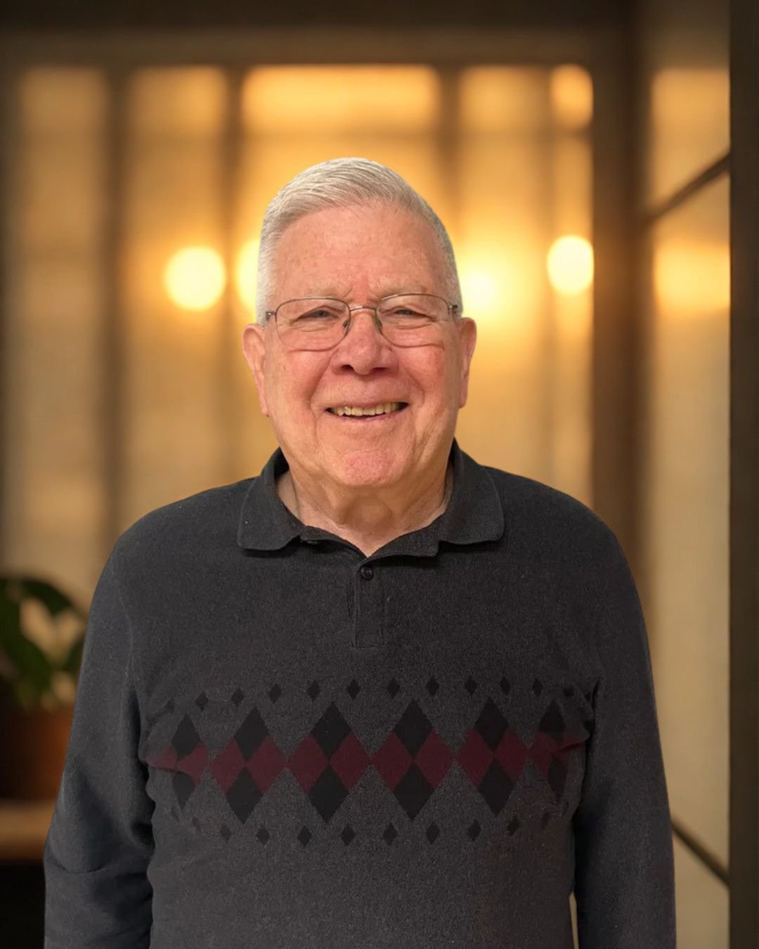 An elderly man with glasses with gray hair, smiling, wearing a black sweater with a diamond pattern on a warm illuminated background.