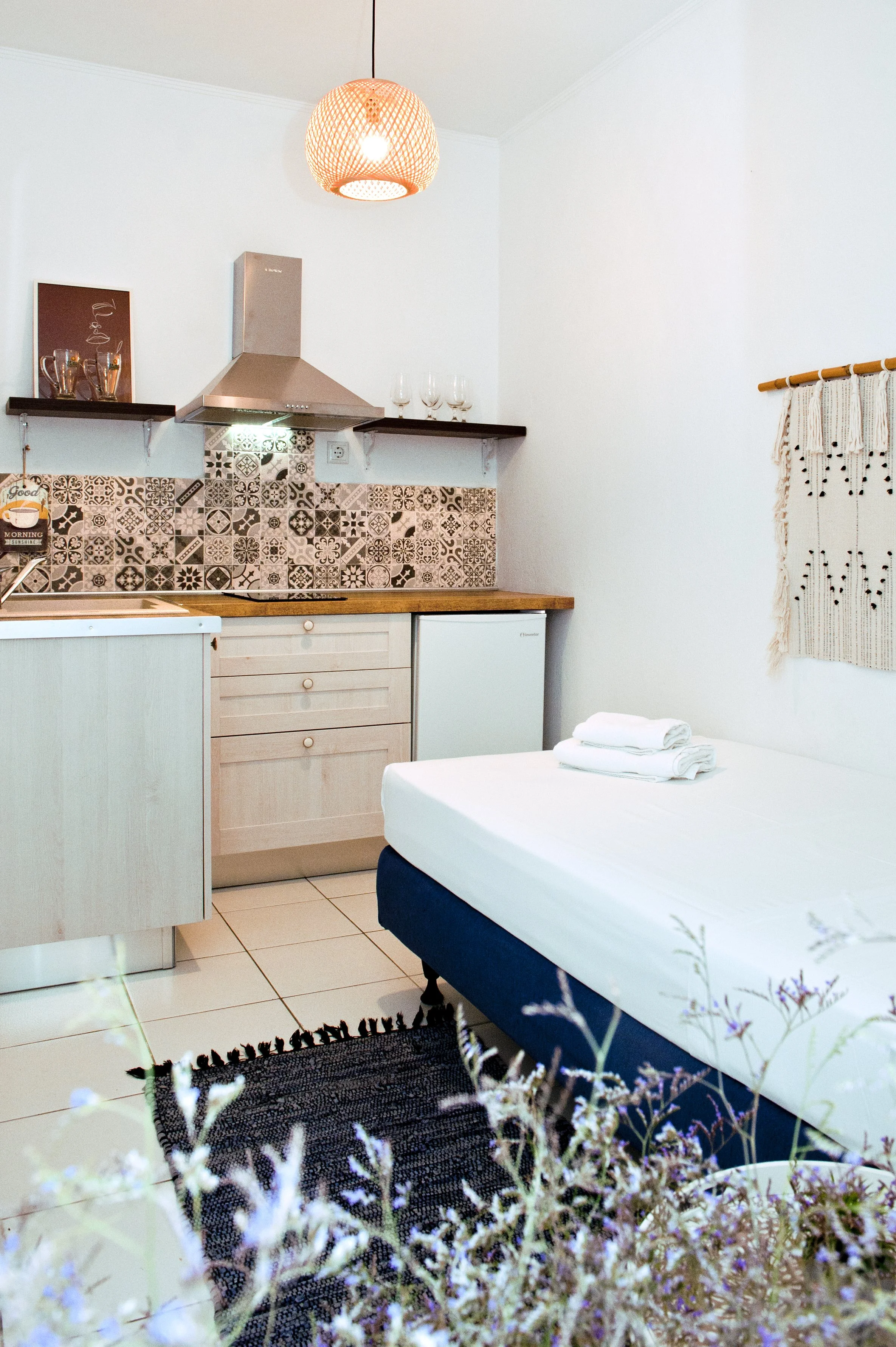 A cozy kitchen area with a white wall, patterned tile backsplash, wooden countertop, and small refrigerator. There are black shelves with glasses and decorative items, a hanging orange lampshade, and a bed with white sheets and folded towels nearby. 