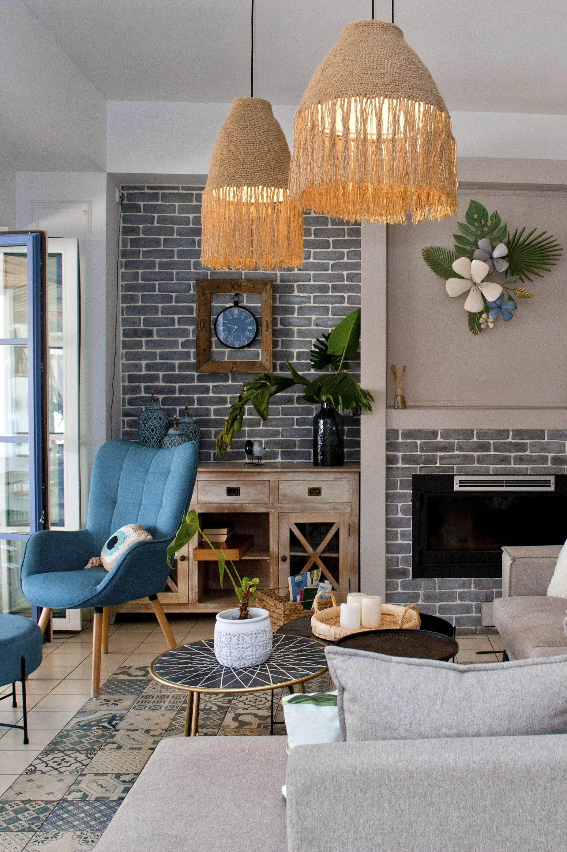 Living room with two woven pendant lights, a brick wall, a wooden console table with decorative items, a blue armchair, plants, a patterned rug, a coffee table with candles, and a wall decor of flowers and leaves.