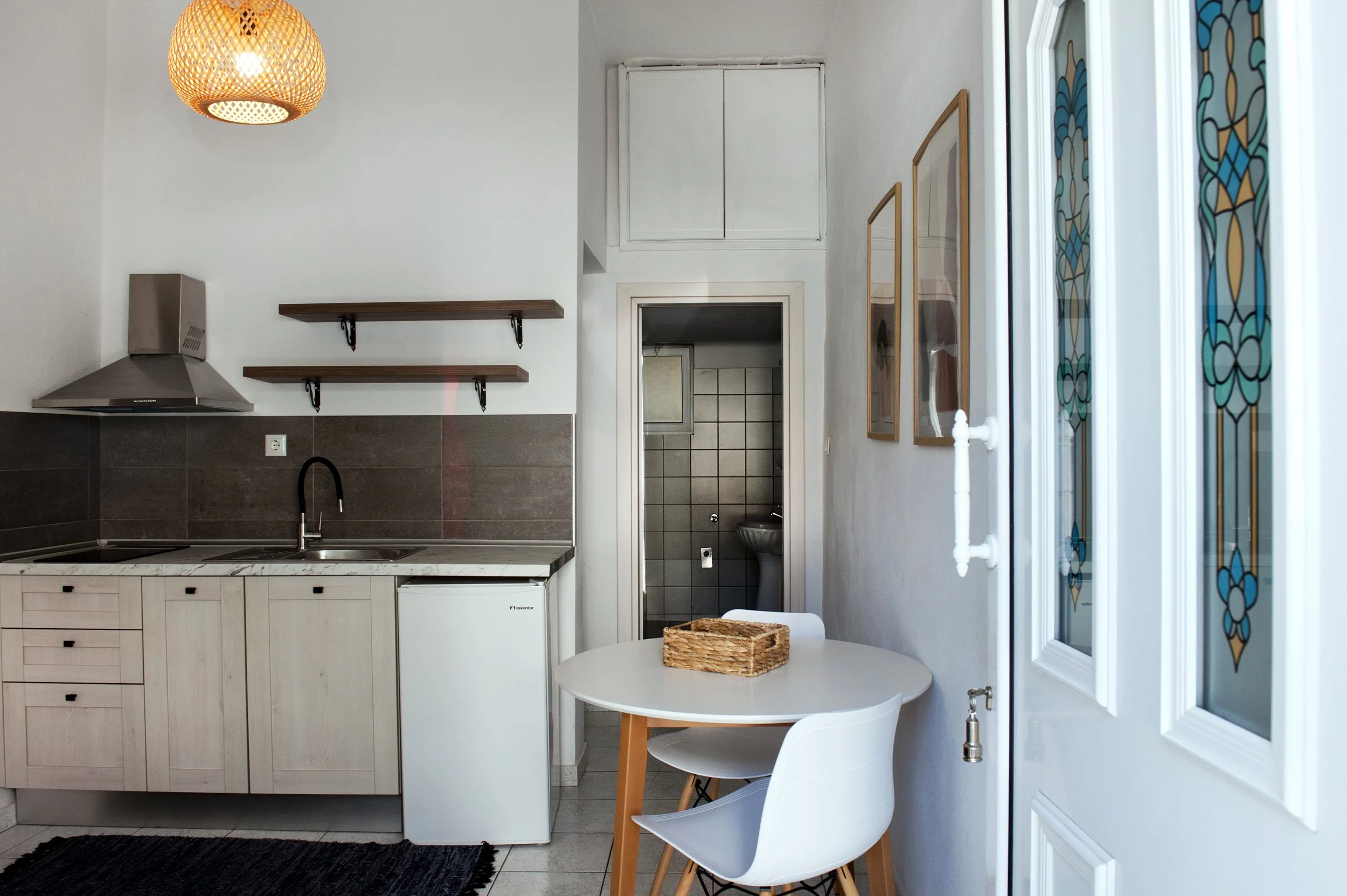 A kitchen with a white countertop, light wood cabinets, two wooden shelves, a grey tiled backsplash, a metallic range hood, a small white refrigerator, and an open door leading to a bathroom with a bidet. There is also a round white table with two wh