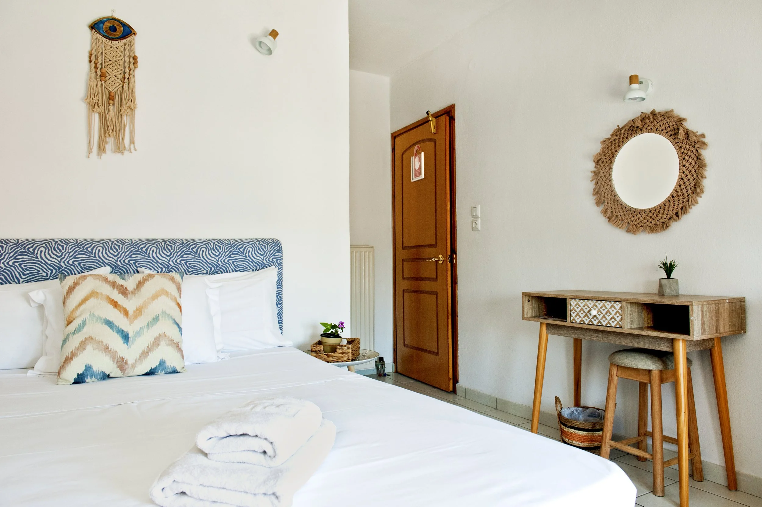 A cozy bedroom corner with a bed decorated with colorful, abstract-patterned pillows. Beside the bed is a small plant on a wicker tray. The wall has a decorative macrame piece and a mirror with a woven frame. There is a wooden table and stool, and a 
