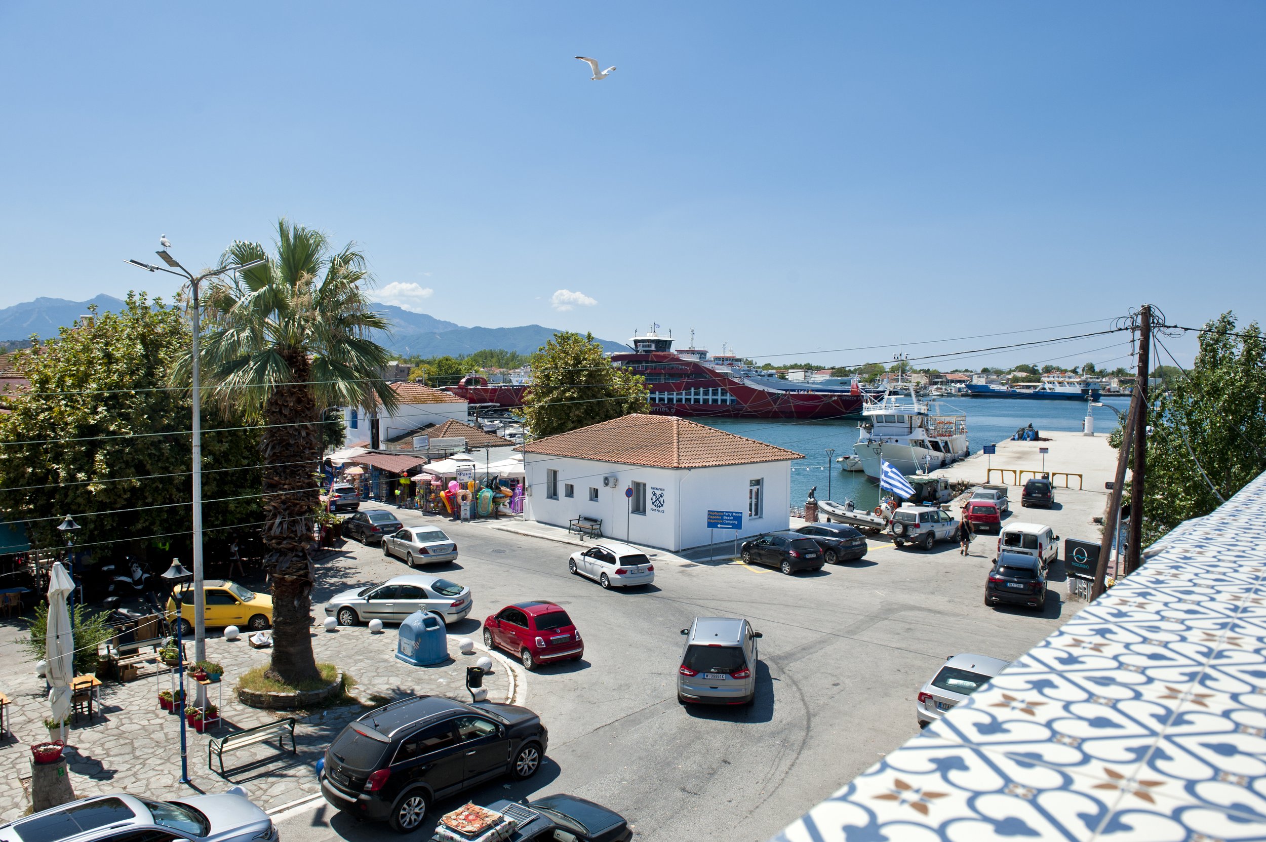 View of a harbor with boats docked, a red ferry, and mountains in the background. A palm tree and a busy parking lot with cars are in the foreground. The sky is clear and blue.