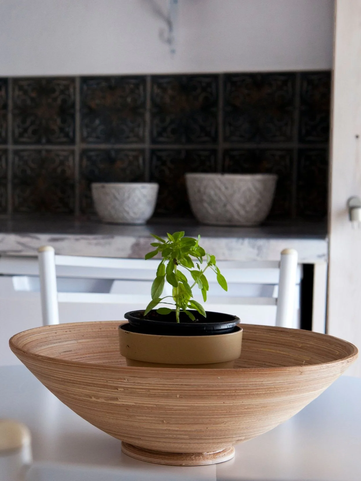 A small green plant in a black pot sits on a round wooden tray on a white table. In the background, two decorative white bowls are on a shelf with a dark, ornate tile backsplash.