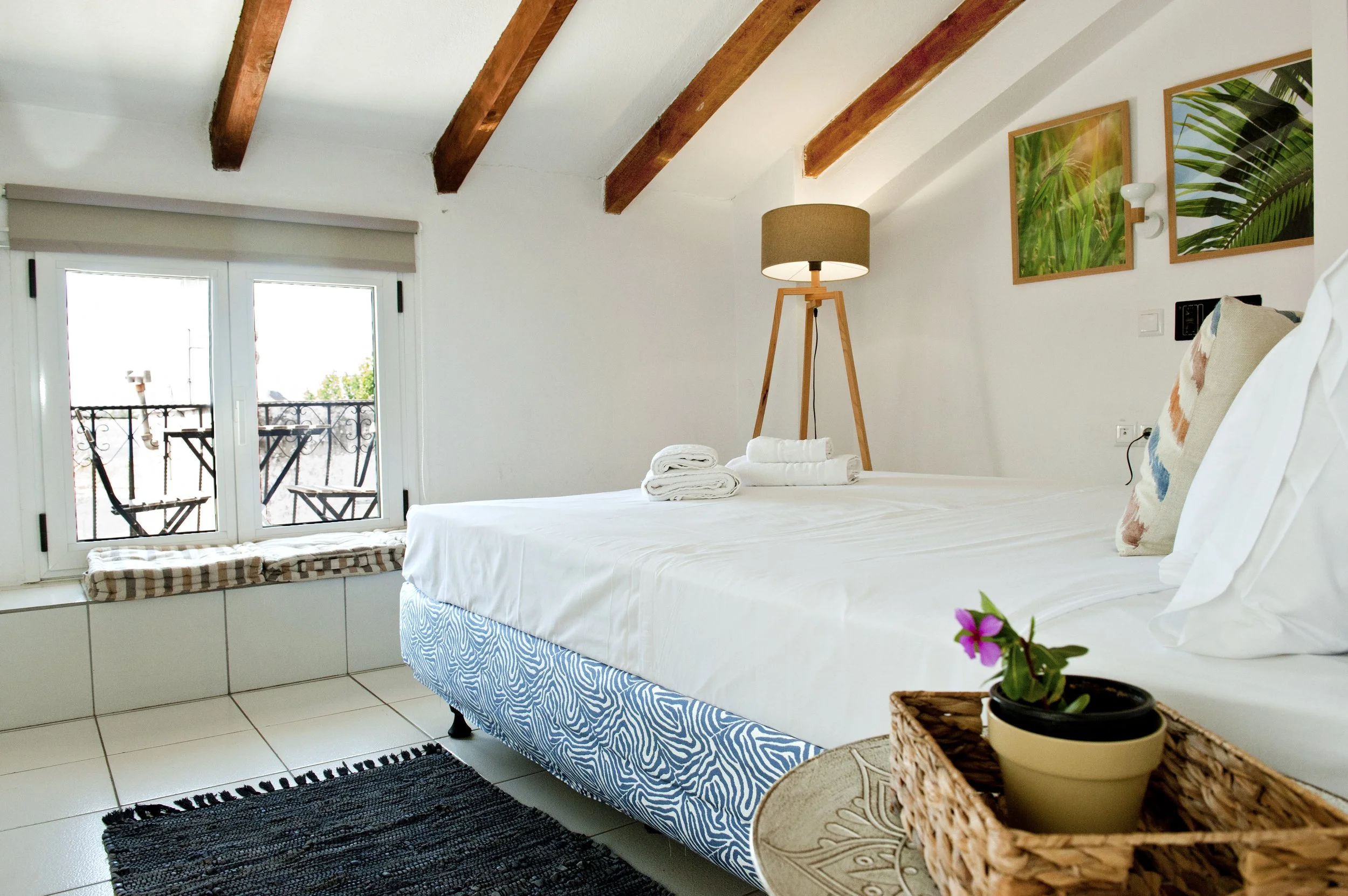 A cozy bedroom with a large white bed, neatly made, with rolled towels on top. There is a white windowed door leading to a small balcony with a black iron railing and chairs. The room has wooden ceiling beams, a floor lamp with a tan shade, framed tr