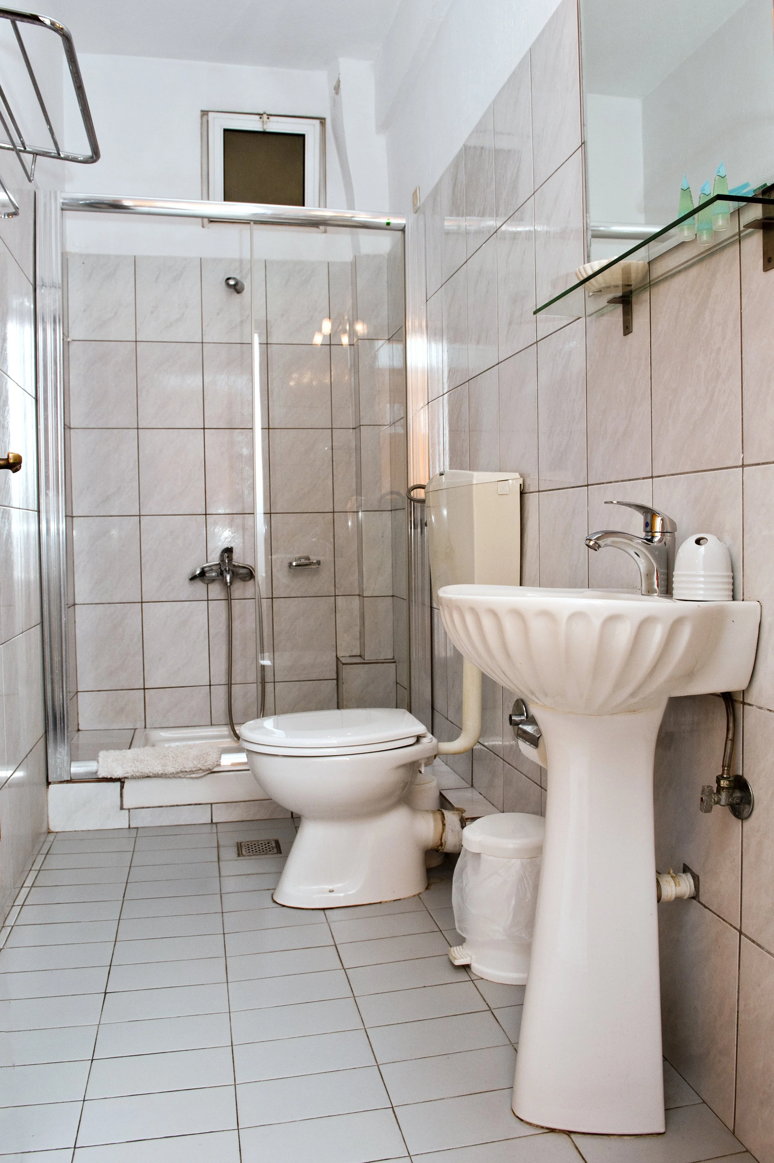 Small bathroom with a toilet, sink, shower enclosure, and a small window. Beige tiles on the walls and floor, with a glass shelf holding toiletries.
