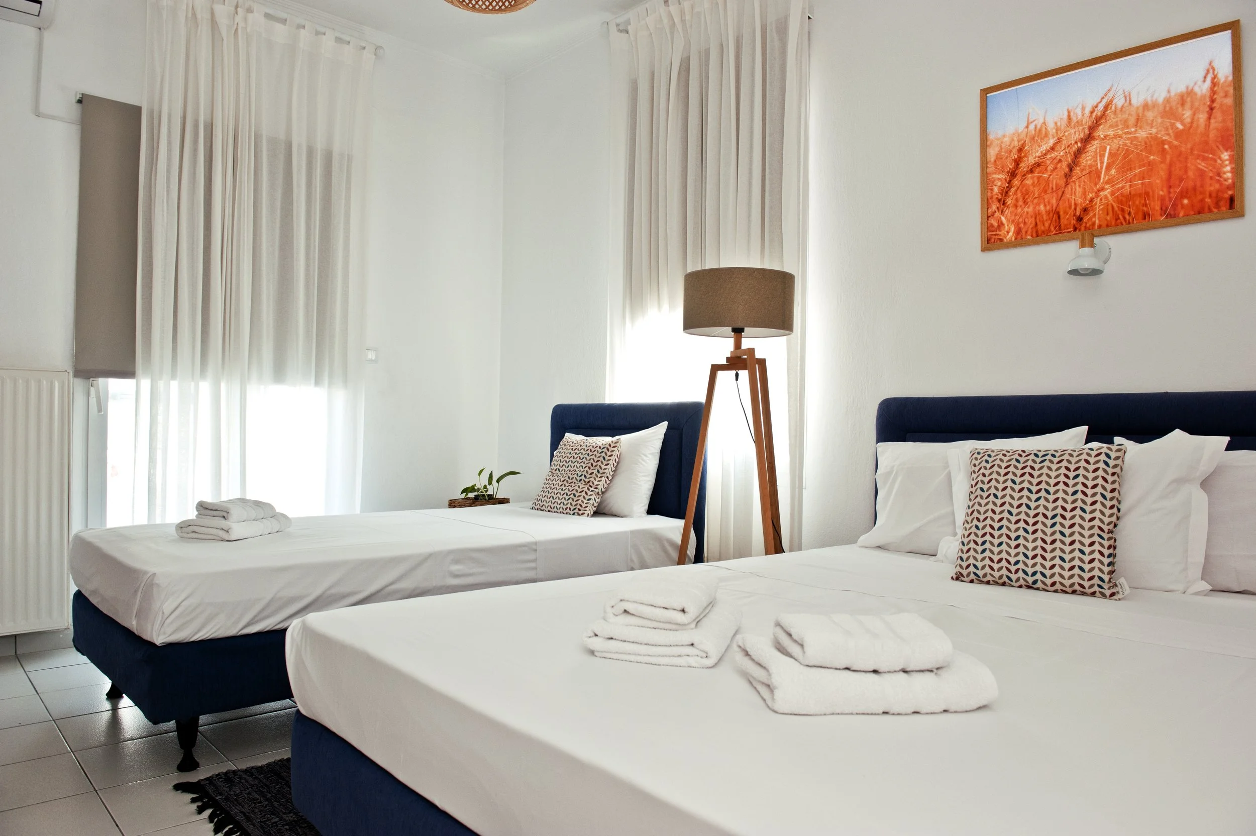 Room with two beds made with white linens, each with folded towels and patterned pillows, a wooden floor lamp, white curtains covering large windows, and a framed picture of orange wheat on the wall.