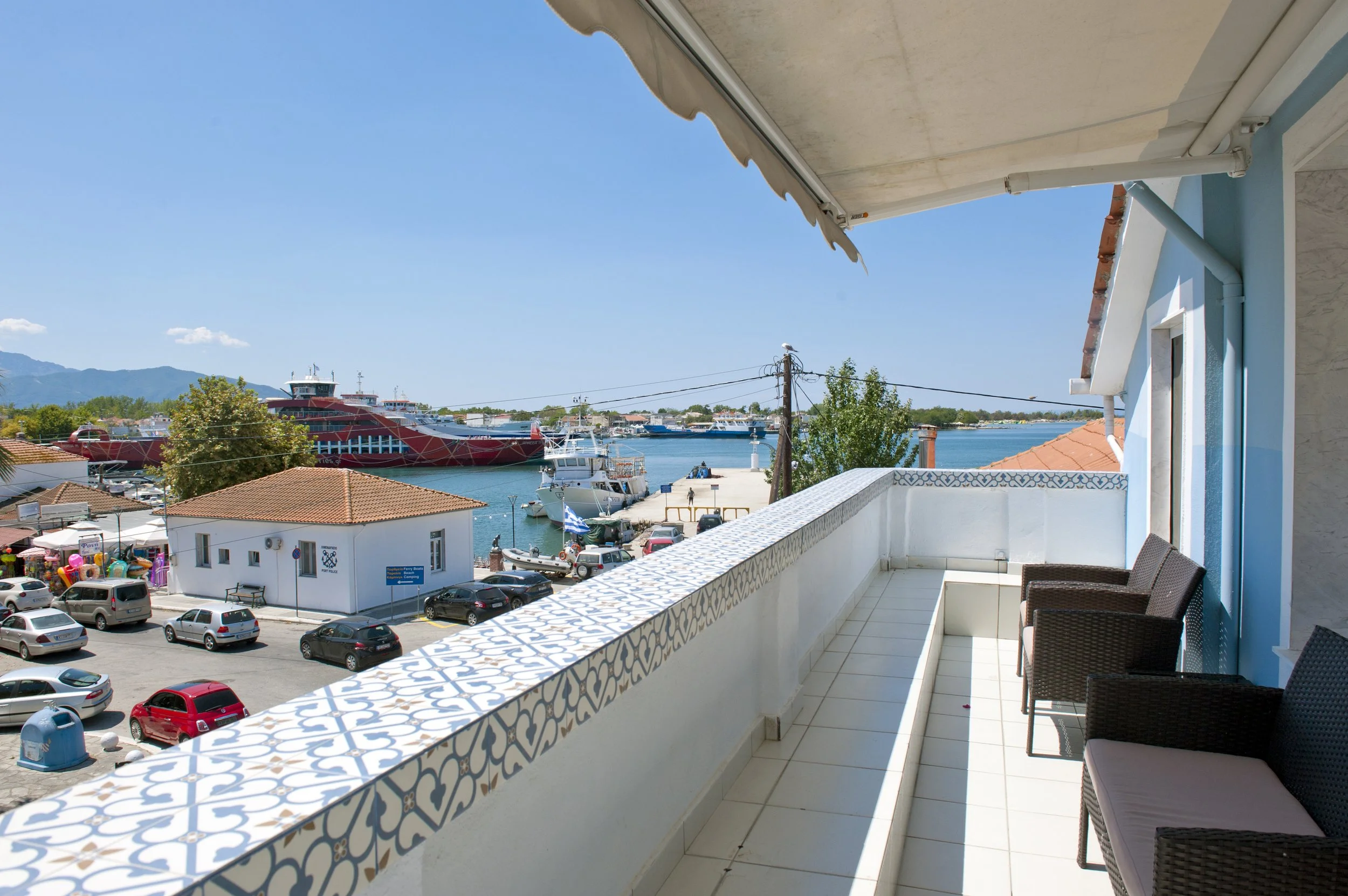 View from a balcony overlooking a marina with boats and a large red cruise ship, with a parking lot below and mountains in the distance under a clear blue sky.