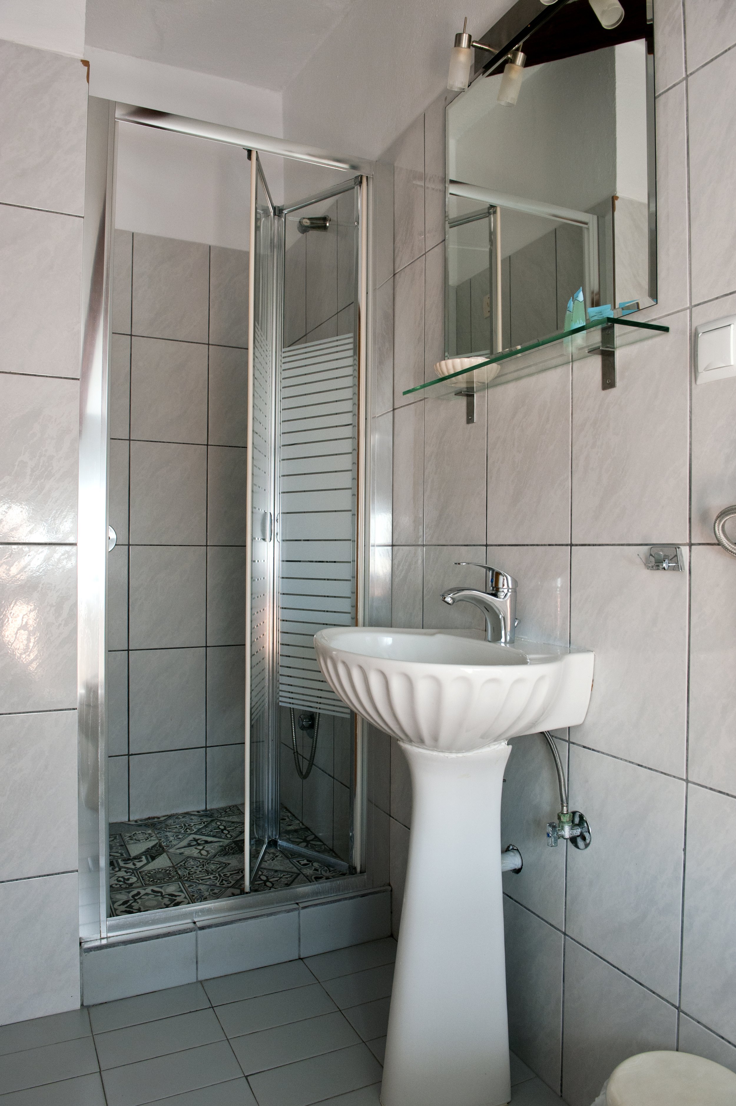 A small bathroom with a white pedestal sink, a rectangular mirror with two lights, and a shower with a glass door and patterned tiles.