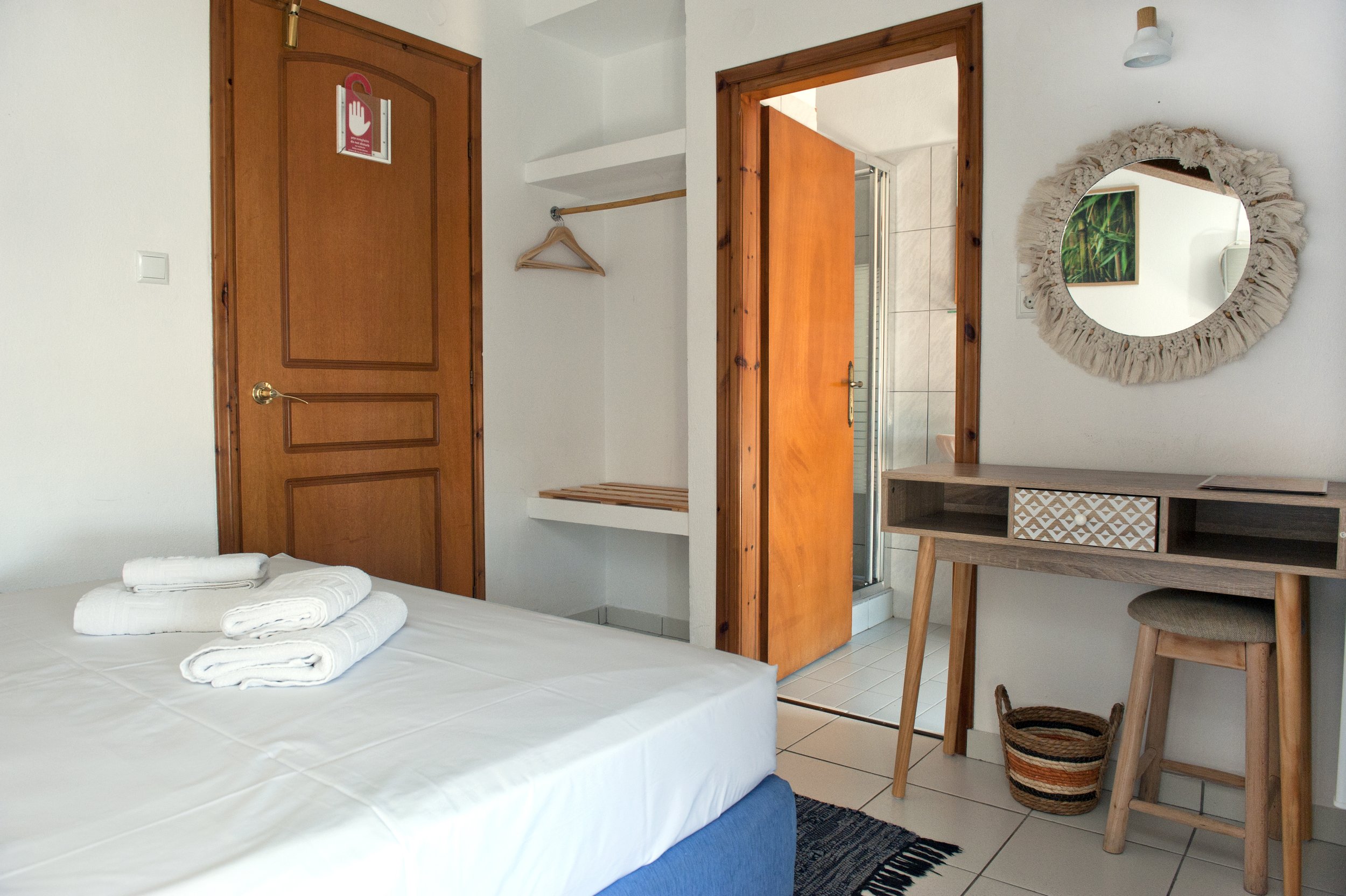 Hotel room with a bed, towels, wooden furniture, a mirror with a woven frame, and a door leading to a bathroom.