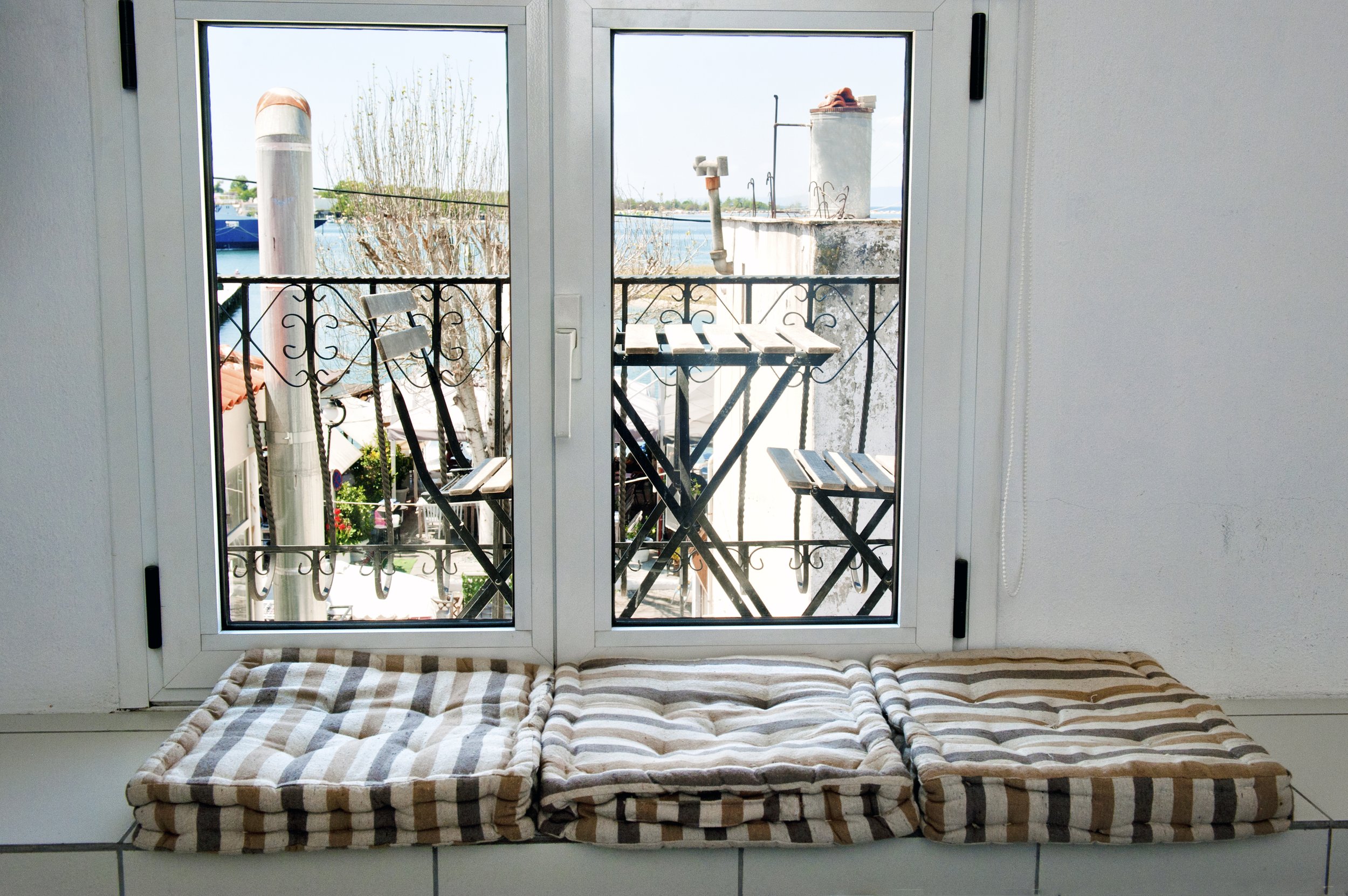 View through a sliding glass door to a balcony with a small round table and chairs, overlooking a body of water with boats and some buildings in the distance.