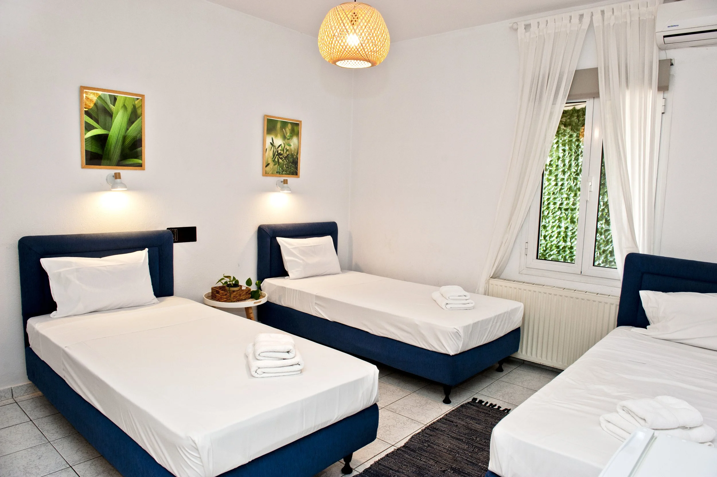 A room with four single beds with white sheets and pillows, two on the left and two on the right, each with a small towel placed on top. There are two framed pictures of greenery on the white wall and wall-mounted lamps above each bed. A window with 