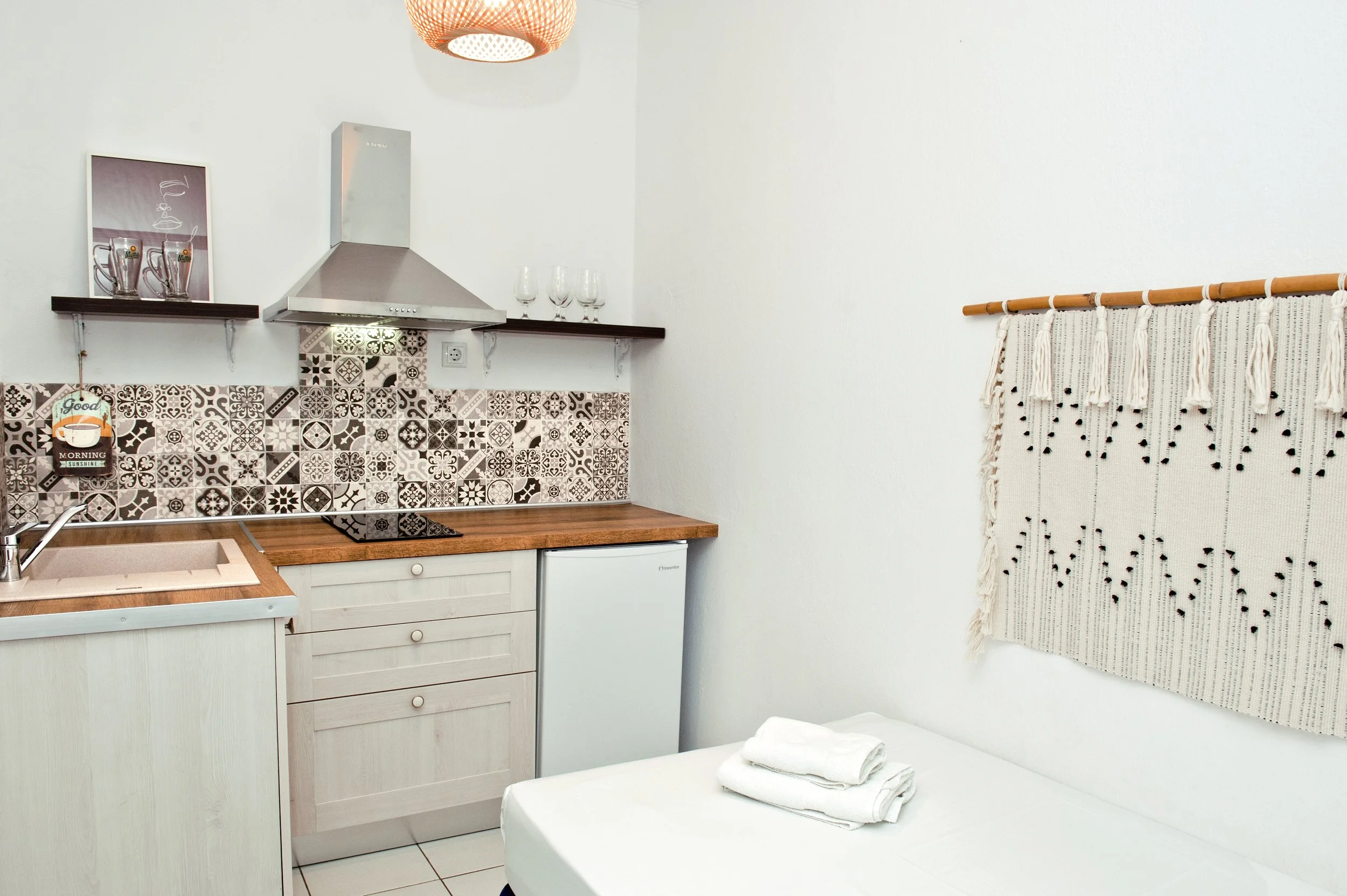 A small kitchen with a patterned tiled backsplash, light wooden cabinets, a countertop with a sink, a mini fridge, and open shelving with glasses. There is a decorative cloth with tassels on the right wall, a light fixture on the ceiling, and a bed w