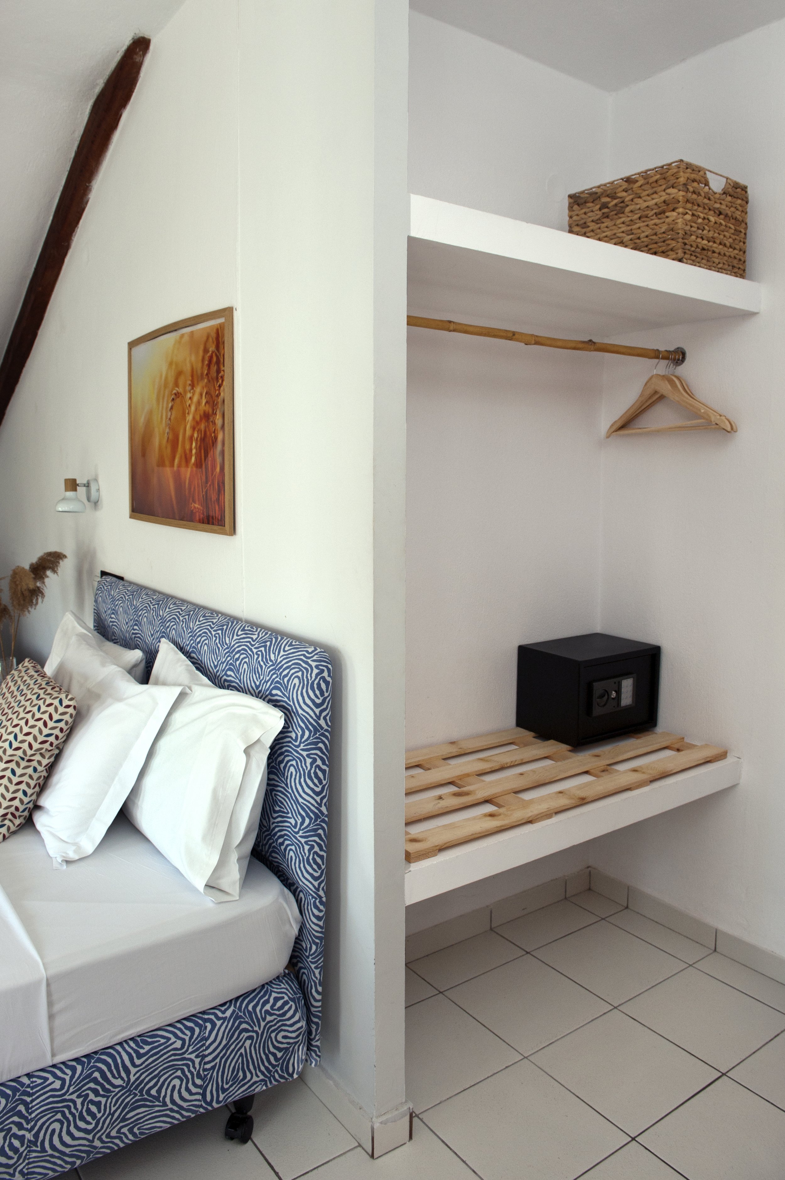 Part of a bedroom with a bed and pillows on the left, and a small empty shelf with a hanging rod and a cardboard box on top on the right.