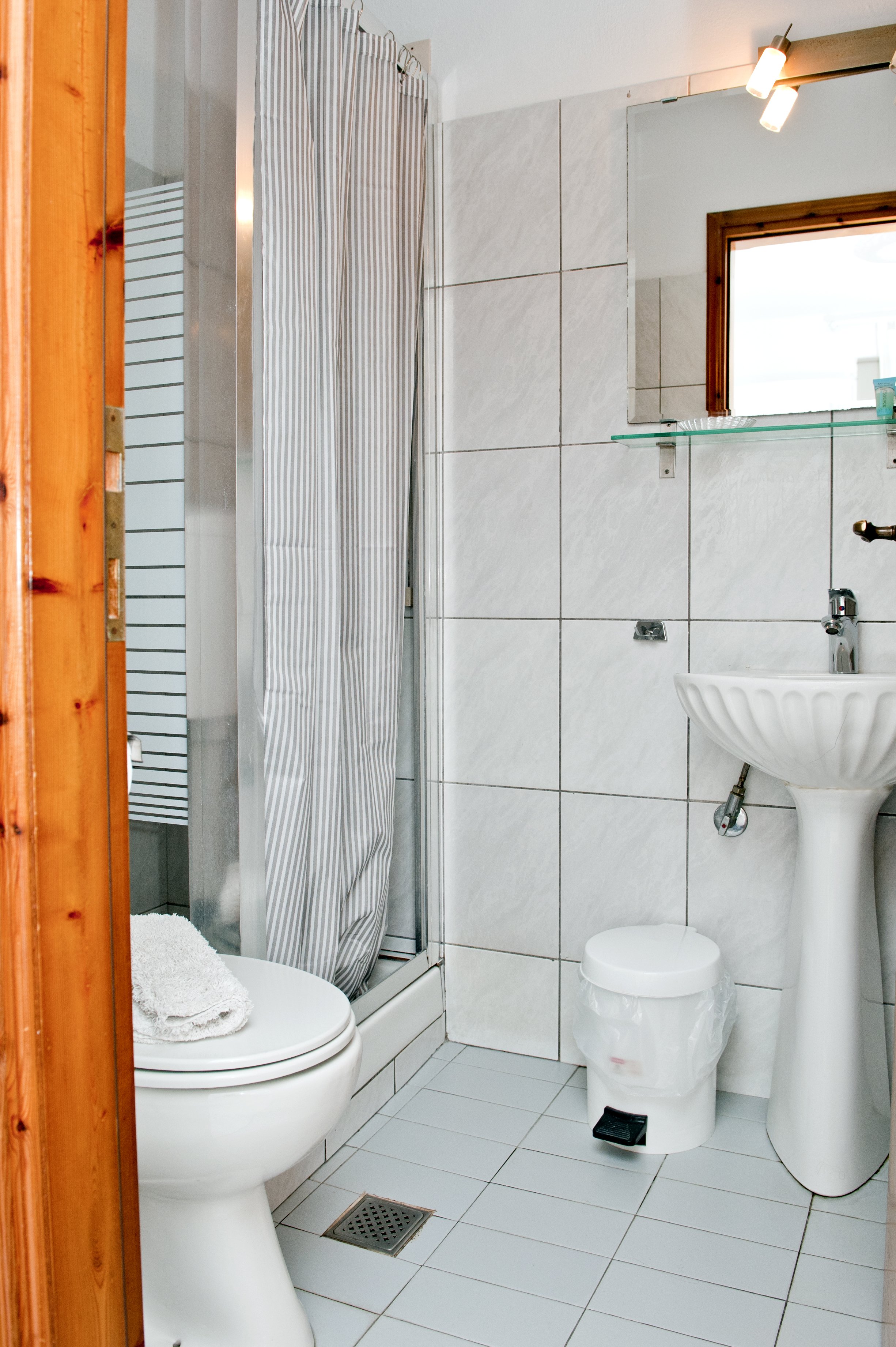 A small bathroom with white tiled walls and floor. There is a toilet with a toilet paper roll on top, and a sink with a round bowl. A shower with a striped curtain is visible on the left. A glass shelf with some toiletries is mounted on the wall abov