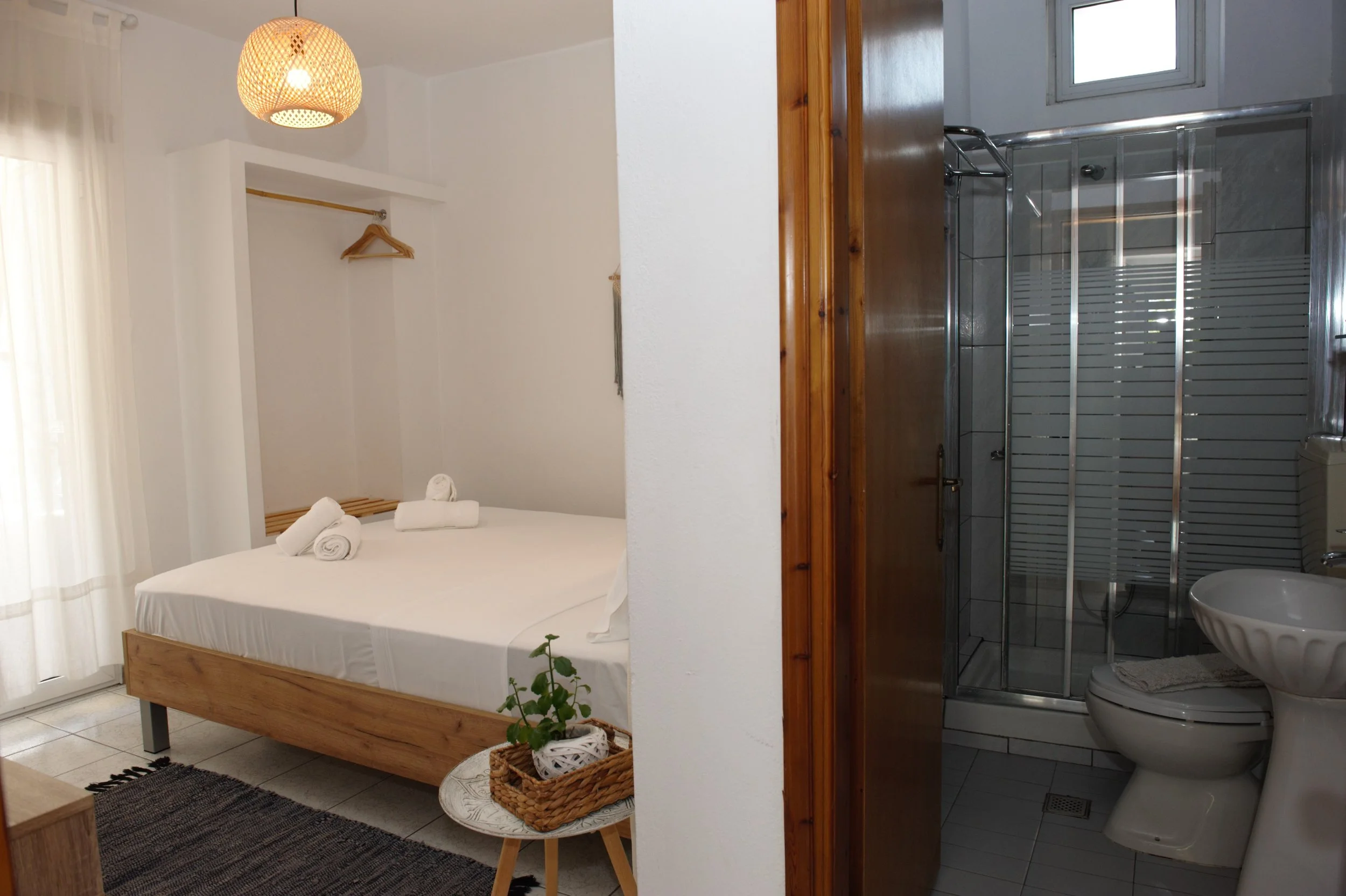 A photo of a cozy bedroom with a wooden bed, white bedding, and rolled towels on top. To the left, a window with sheer white curtains lets in natural light, and a woven pendant light hangs from the ceiling. There is a small wooden side table with a p