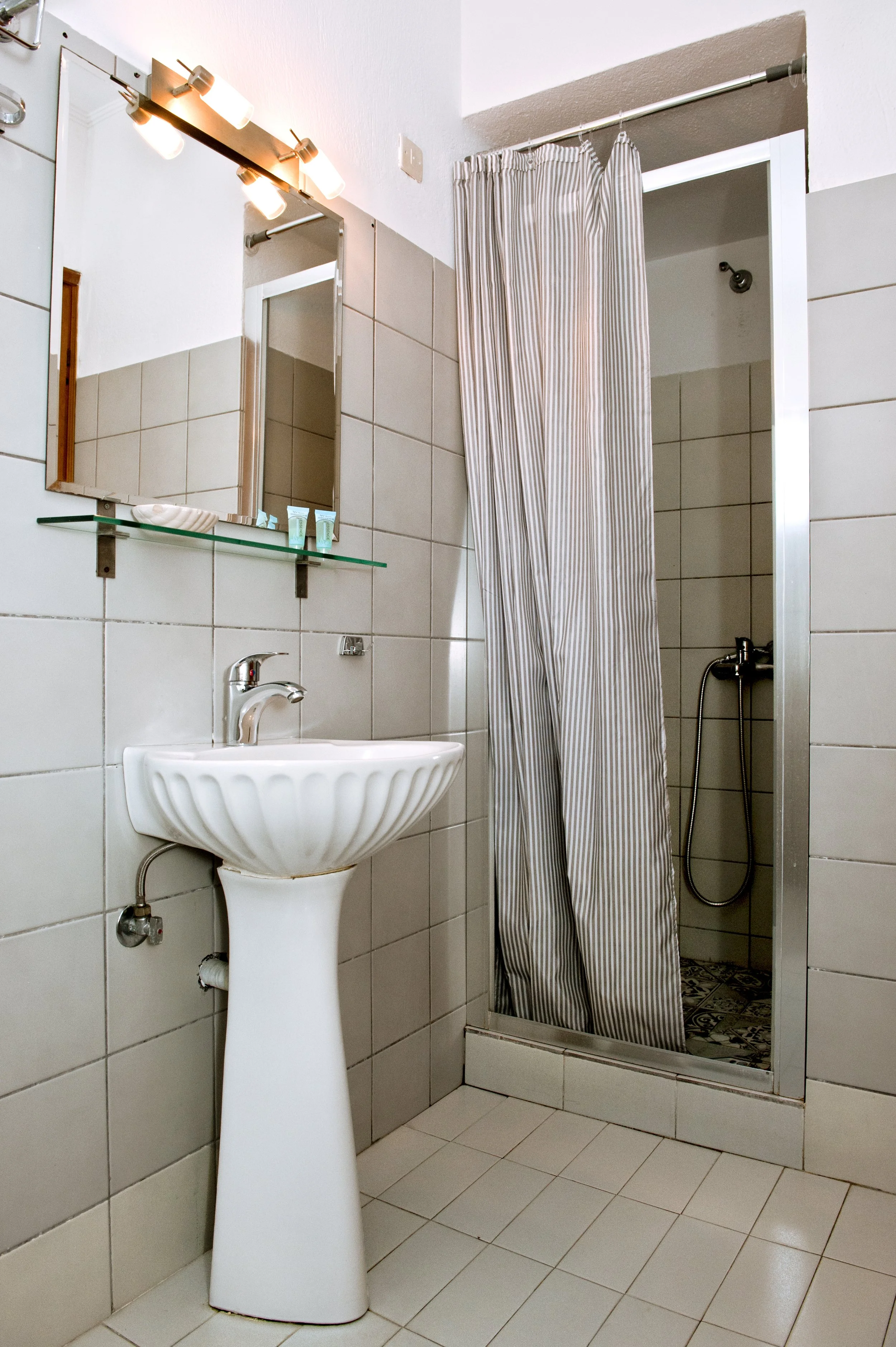 Bathroom with a white pedestal sink, a mirror, a glass shelf with toiletries, and a shower area with beige and white striped curtain and patterned floor tiles.