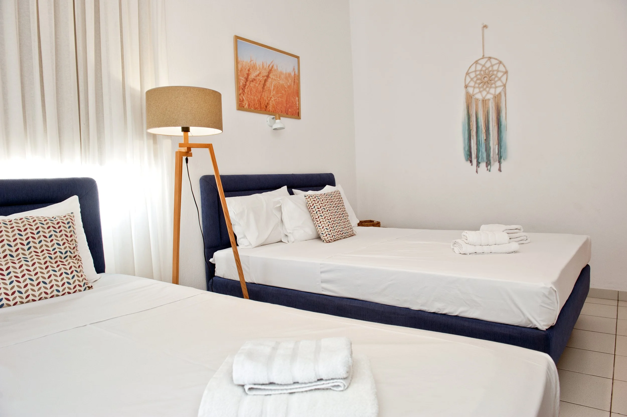 A bright bedroom with two beds, each with patterned pillows, a bedside lamp, a picture of a wheat field, a dreamcatcher, and neatly folded towels.