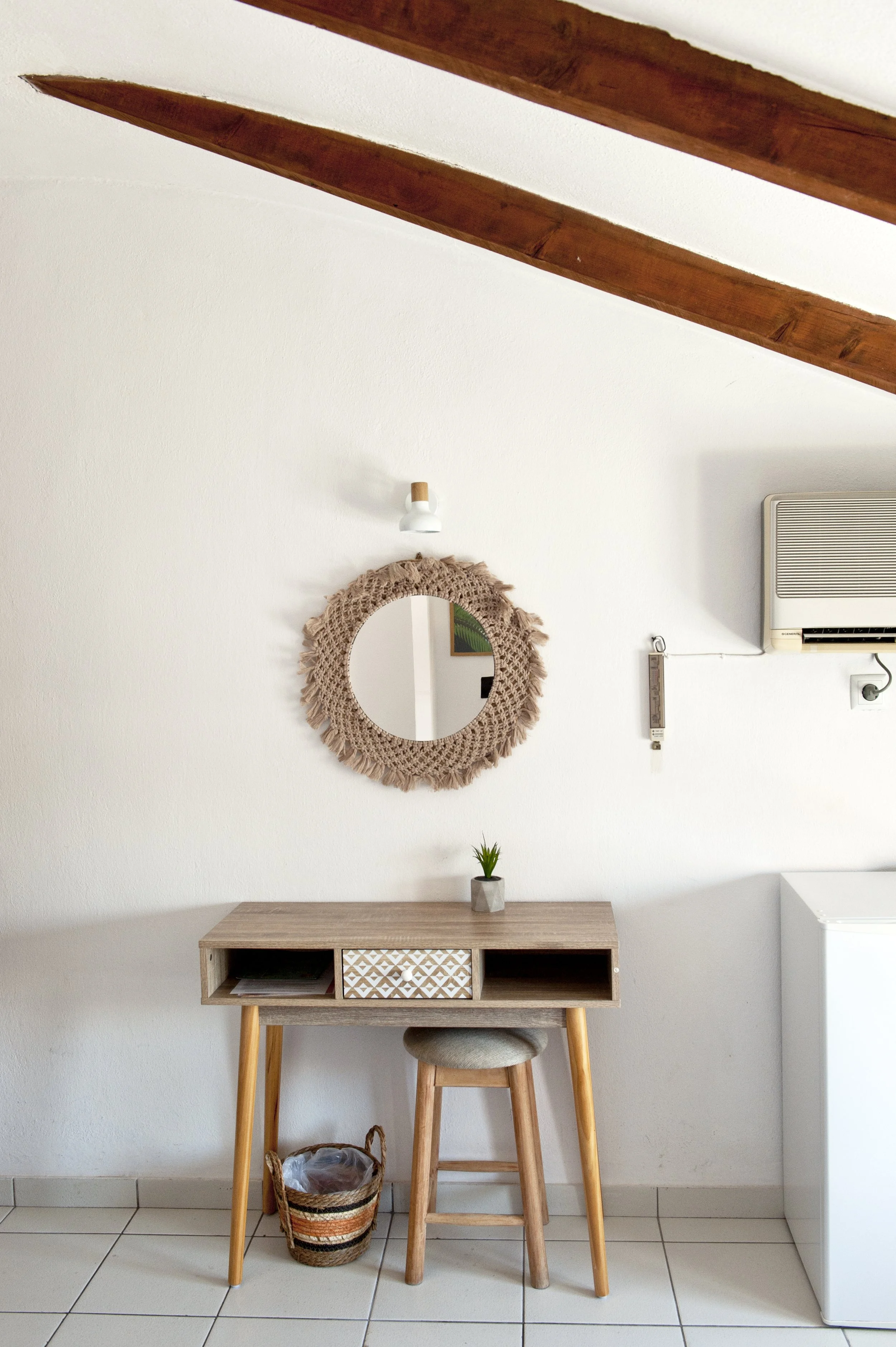 A small wooden desk with a drawer unit and open space beneath it, a wooden stool with a cushioned top, a woven basket on the floor, a mirror with a woven frame hanging on a white wall, and an air conditioning unit mounted high on the wall in a minima