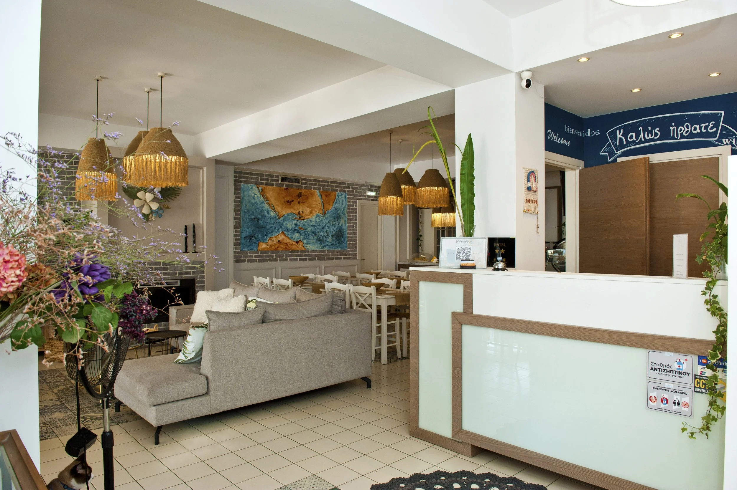 Interior of a hotel lobby with a reception desk, beige sofa, dining area with wooden chairs, colorful abstract wall art, hanging pendant lights with fringe, plants, and floral arrangements.