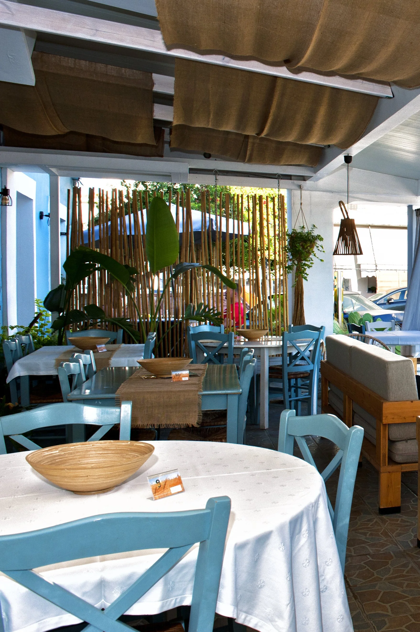 Interior of a restaurant or cafe with blue chairs and tables covered with white tablecloths, a wooden bowl on one table, and decorative plants and bamboo divider in the background.