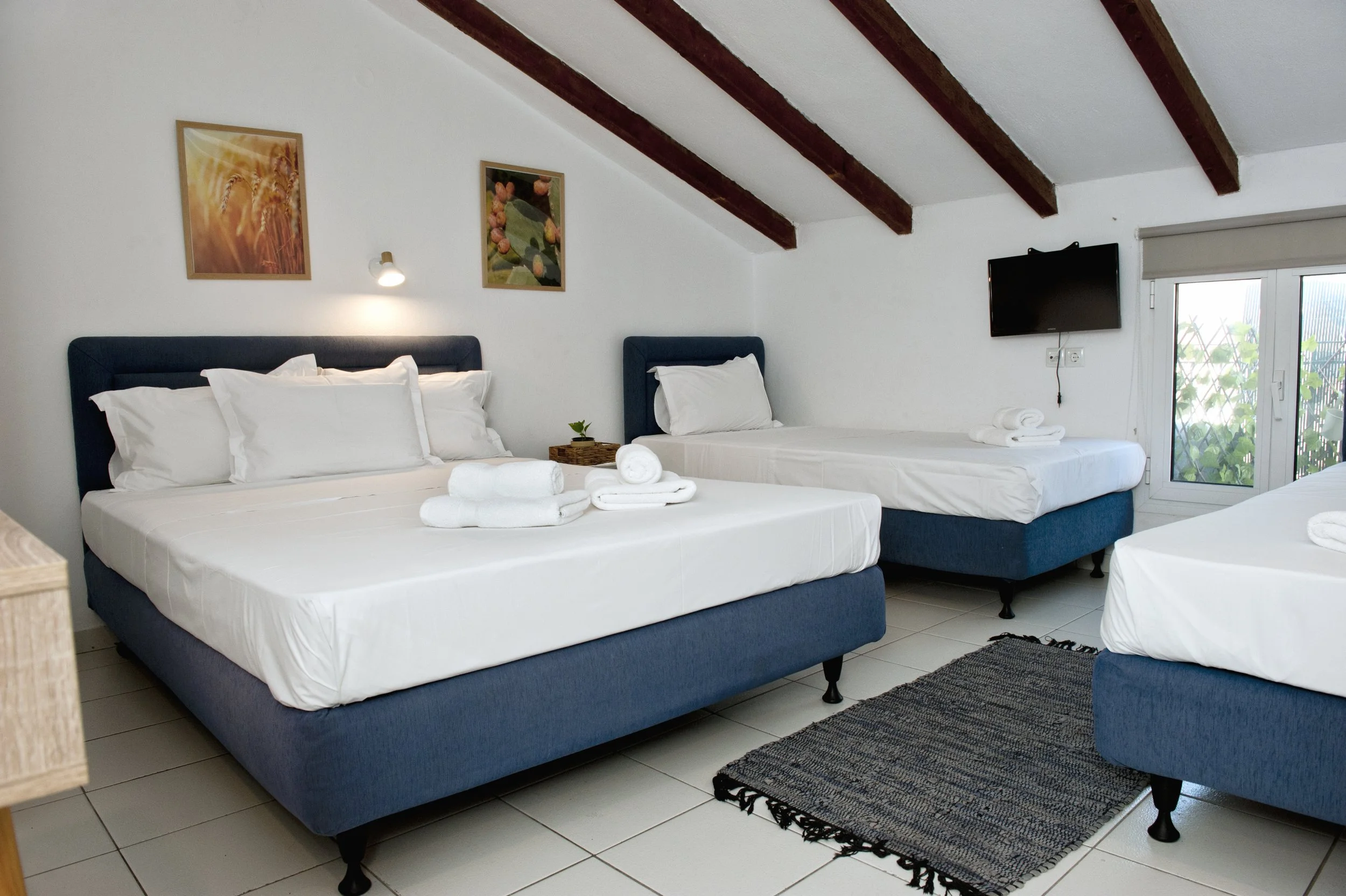 A bright, cozy bedroom with three beds featuring navy blue upholstered headboards and white bedding. The room has white walls with two framed pictures and a small wall lamp above the beds. Natural light comes through a window with a roller shade, and